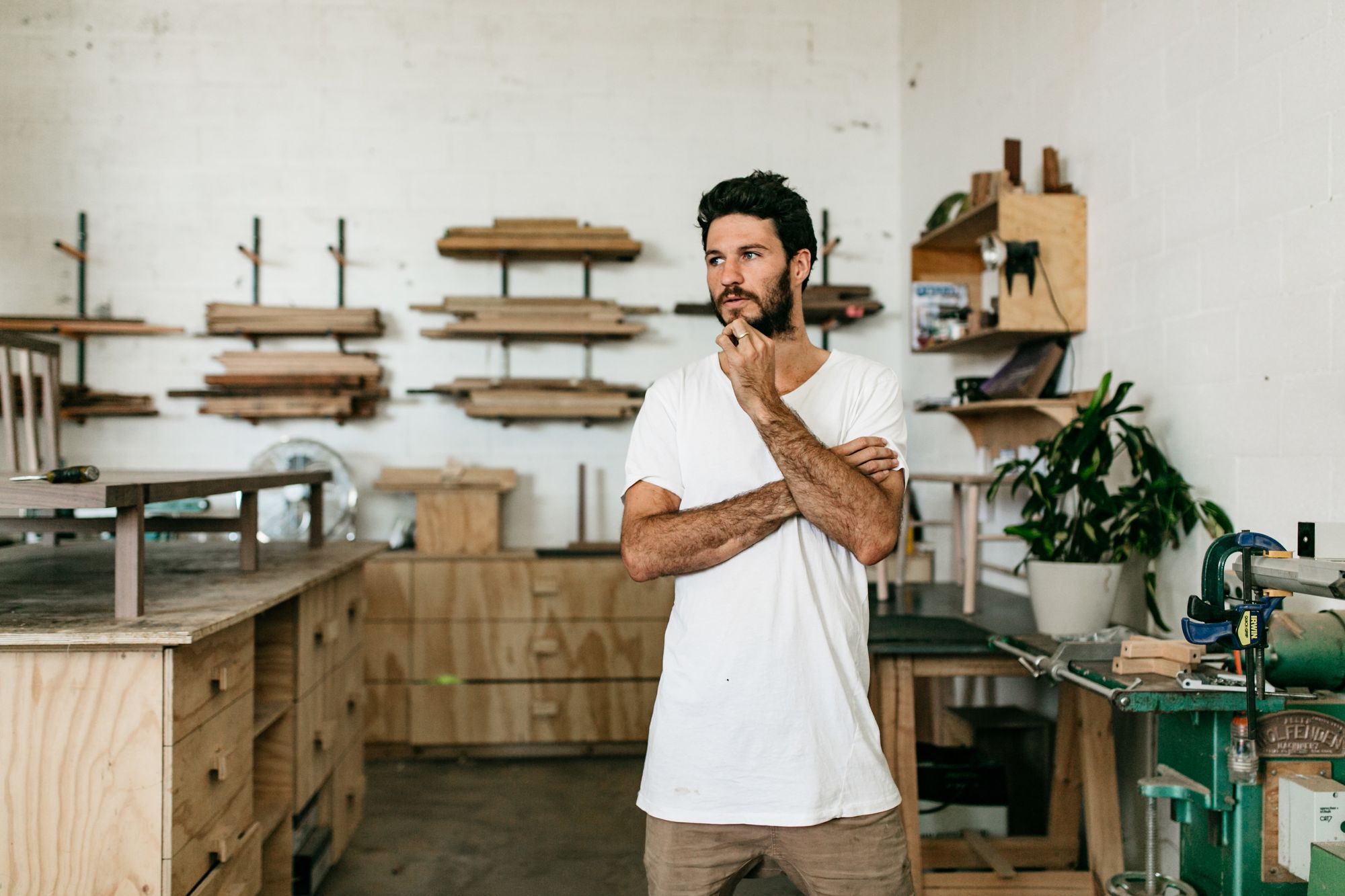 A man in a white t-shirt stands contemplatively in a woodworking workshop. He has one hand raised to his chin, deep in thought. The workshop is equipped with various wooden shelves containing planks of different sizes, a table with a vice, and a few other carpentry tools. The walls are painted white, contrasting with the natural colors of the wood and the green of a potted plant.