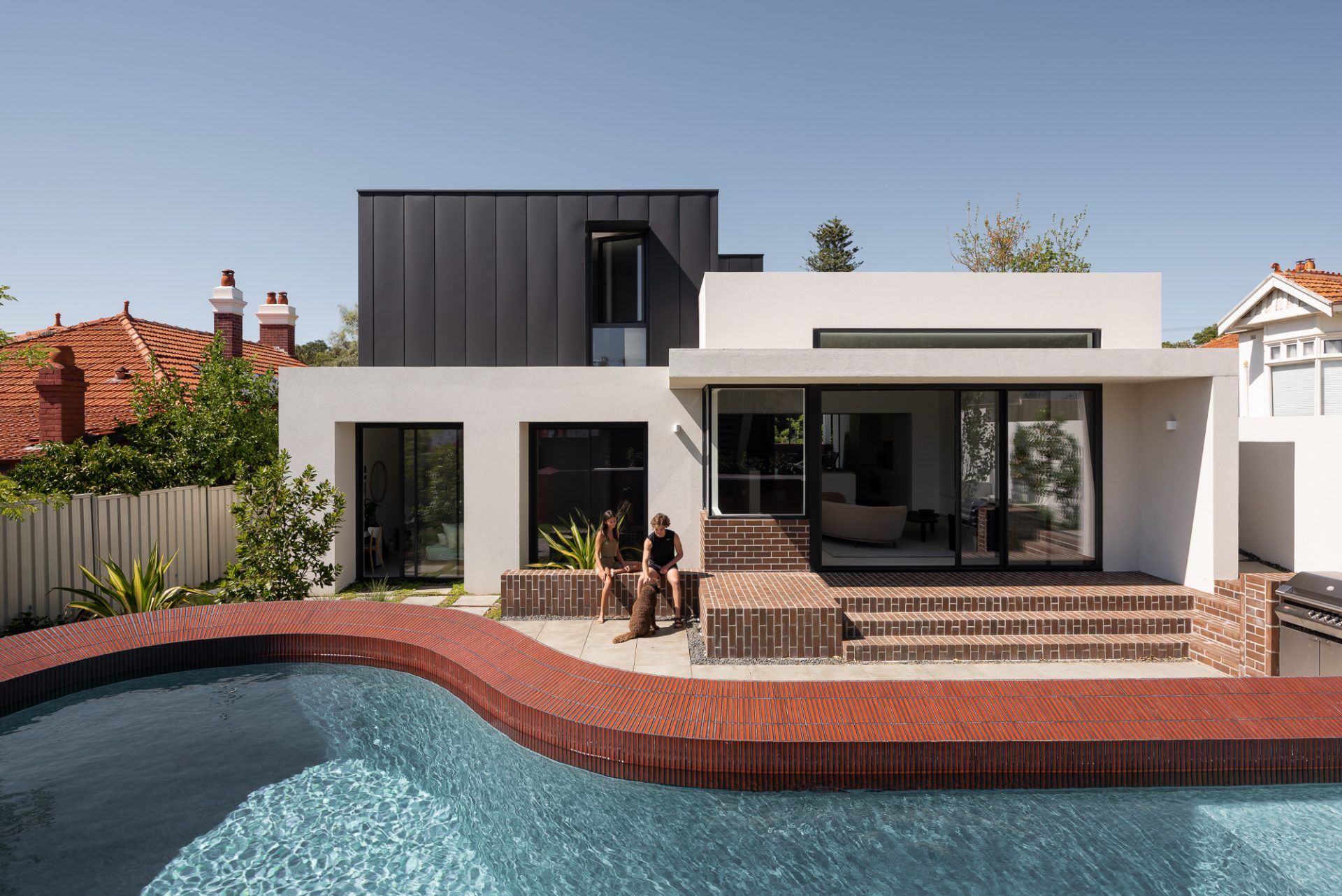 Hyde Park House by Robeson Architects. Rear facade of modern, double story home and raised pool area. Structure built from red brick, white render and the second story comprised of black metal. 