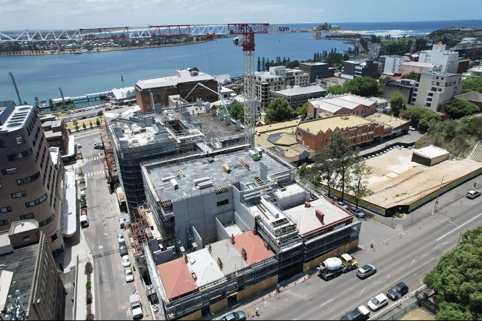 An aerial view showcases an urban construction site amidst a backdrop of diverse architectural structures. Dominating the frame's center is a building under construction, wrapped in scaffolding, with workers busy on its levels. Construction materials and equipment are scattered across the site, and a tall white crane towers above, casting its shadow below. Adjacent to the site are a mix of older brick buildings, modern apartments, and commercial establishments. On the left, a glimpse of a bright red bridge stands out against the blue water of a vast river or bay. Farther in the distance, a coastal line with trees and a beach area can be spotted.