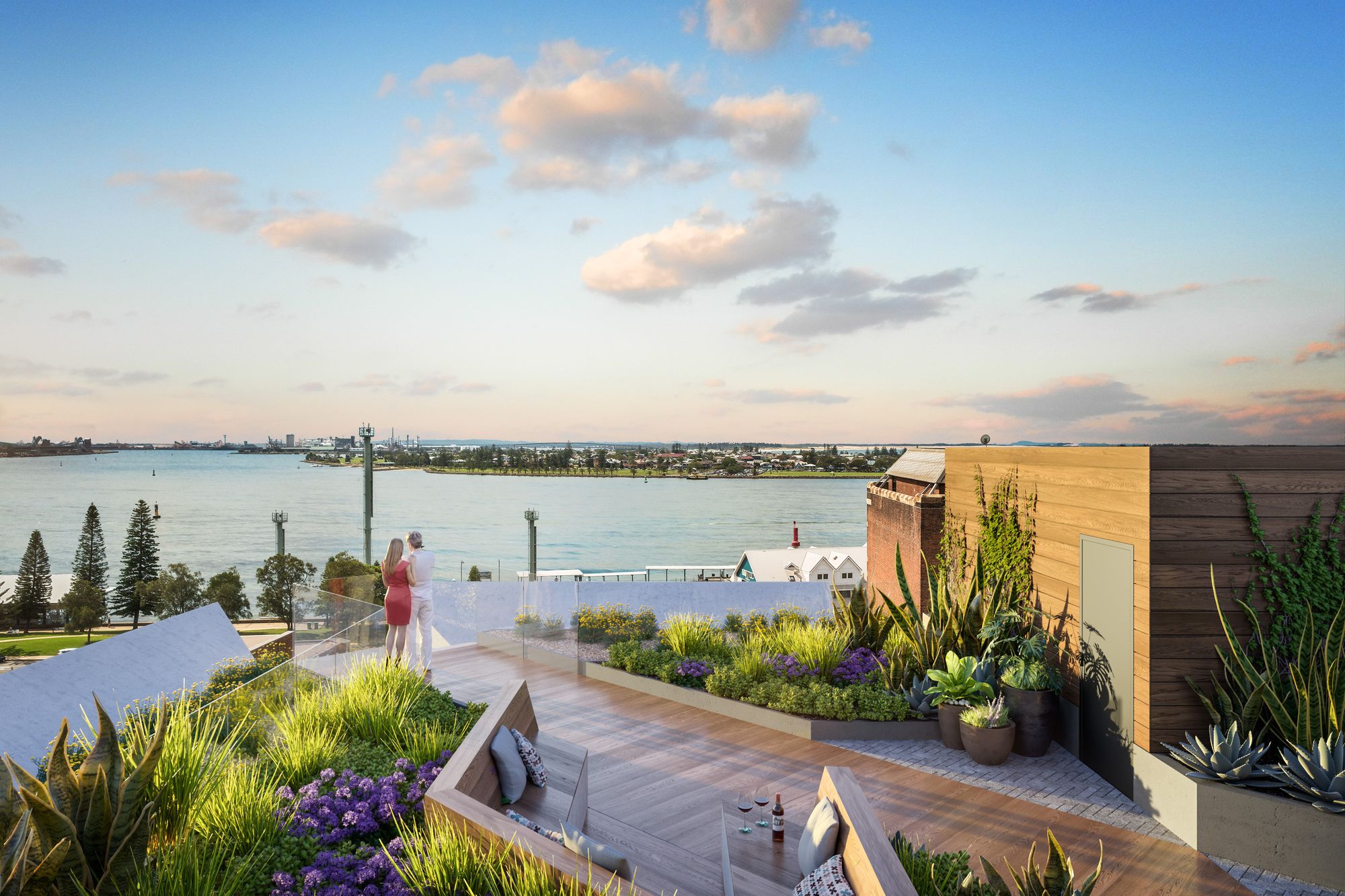 The image showcases a rooftop terrace garden during the evening with a breathtaking view of a serene body of water and a distant city skyline. The terrace is adorned with wooden decking and a variety of lush, well-maintained plants. These include vibrant purple flowers, tall grasses, and succulents. A sunken seating area with plush cushions and a small table holding beverages provides a cozy spot for relaxation. On the right, a wooden structure with climbing plants forms a green wall, and an adjacent brick building adds a touch of historic charm. In the foreground, two individuals stand by a transparent barrier, admiring the picturesque view, bathed in the warm hues of the setting sun. The tranquil atmosphere suggests a perfect blend of urban living and nature's serenity.     Is this conversation helpful so far?