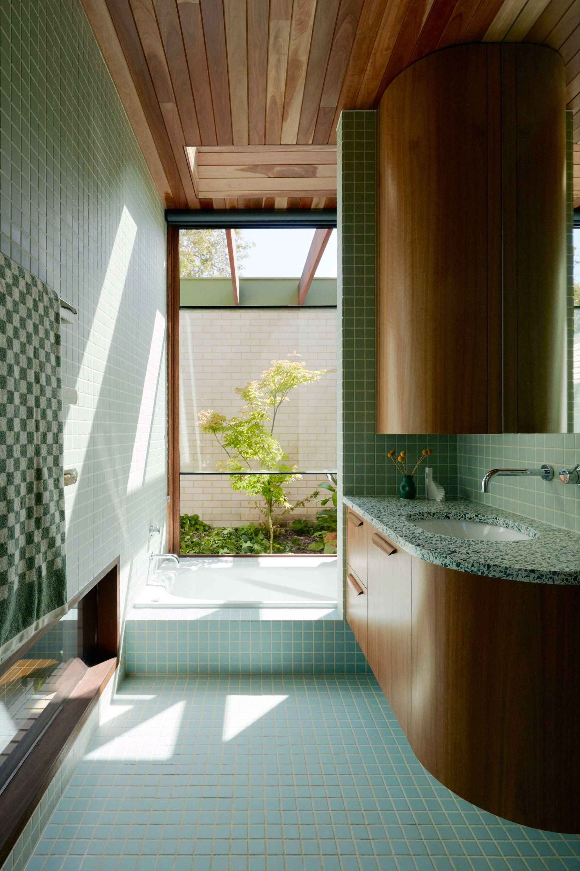 Florida House by Nest Architects with Placement Studio showing a green tile and timber bathroom with a garden outlook