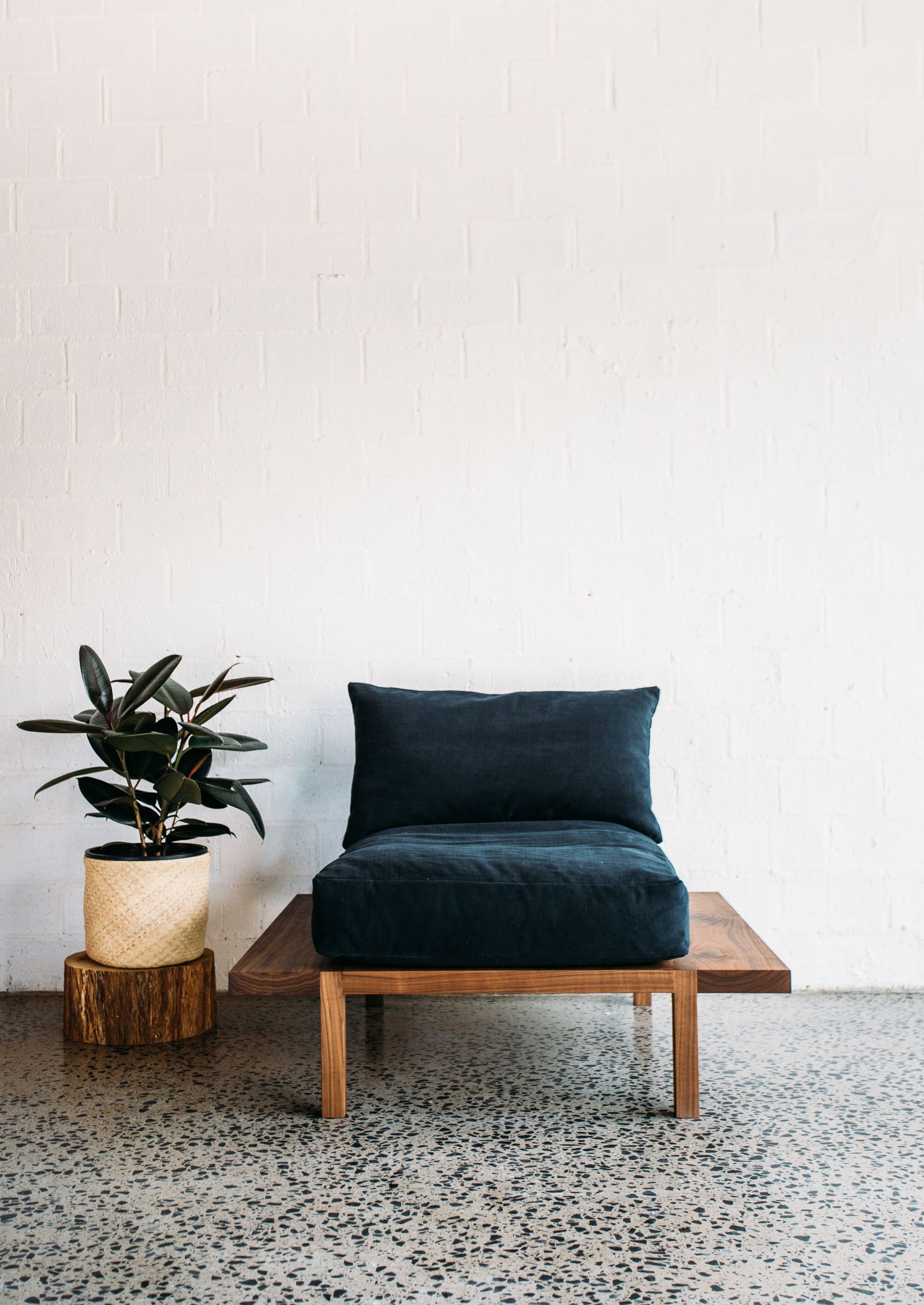 J.D Lee Furniture. A cozy corner featuring a dark charcoal cushioned chair with a wooden base, set against a white brick wall. Beside the chair, there's a green potted plant with dark leaves placed on a wooden stand, which is capped with a woven patterned pot. The scene is completed with a terrazzo-style floor that adds texture to the minimalist setting. The overall aesthetic is modern with a touch of nature and contrasting textures.     User Uploaded image alt text.  ChatGPT A warm and inviting dining setup showcasing a rich wooden table with a smooth finish. Atop the table are ceramic pieces, including a tall cylindrical vase, a brown bottle-shaped container, and a shallow bowl in an earthy hue. The table is accompanied by chairs with matching wooden frames and woven rattan seats, creating a harmonious blend of textures. The terrazzo-style flooring provides a neutral backdrop, complementing the natural and minimalist aesthetic of the scene. The overall ambiance suggests a serene and rustic elegance.     User Uploaded image alt text.  ChatGPT A contemporary wooden lounge chair with a minimalist design sits gracefully against a backdrop of white walls. The chair features a deep seat cushioned with soft white pillows, providing a stark contrast to its rich brown frame. Its backrest consists of evenly spaced wooden slats, adding an element of geometric visual interest. The polished concrete flooring, speckled with unique textures, complements the simple yet elegant aesthetic of the scene. The overall setting exudes a sense of calmness and refined modernity.