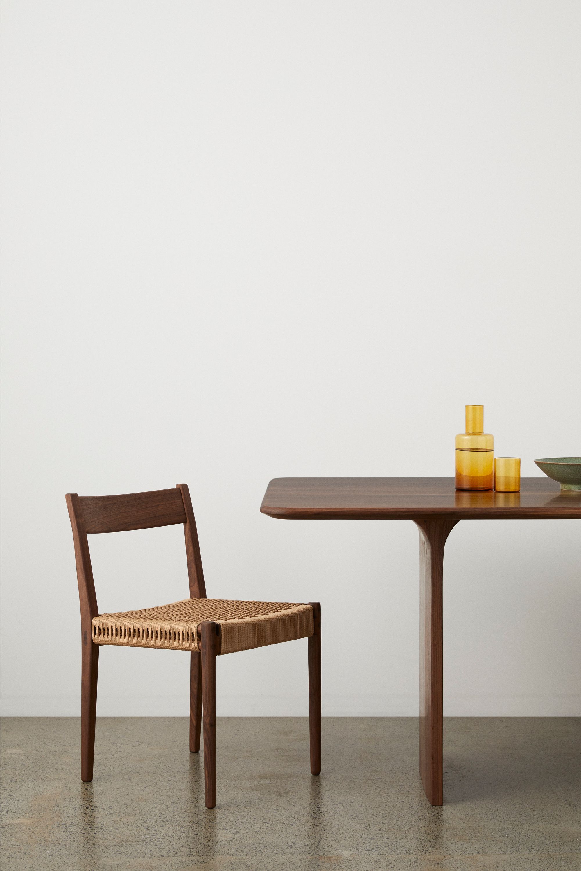 JD. Lee Furniture. A minimalist setting featuring a wooden dining chair with a woven seat, positioned adjacent to a matching wooden table. On the table, there are two yellow-toned glass containers of different sizes and a small greenish bowl. The furniture is set against a plain white backdrop and rests on a speckled concrete floor.