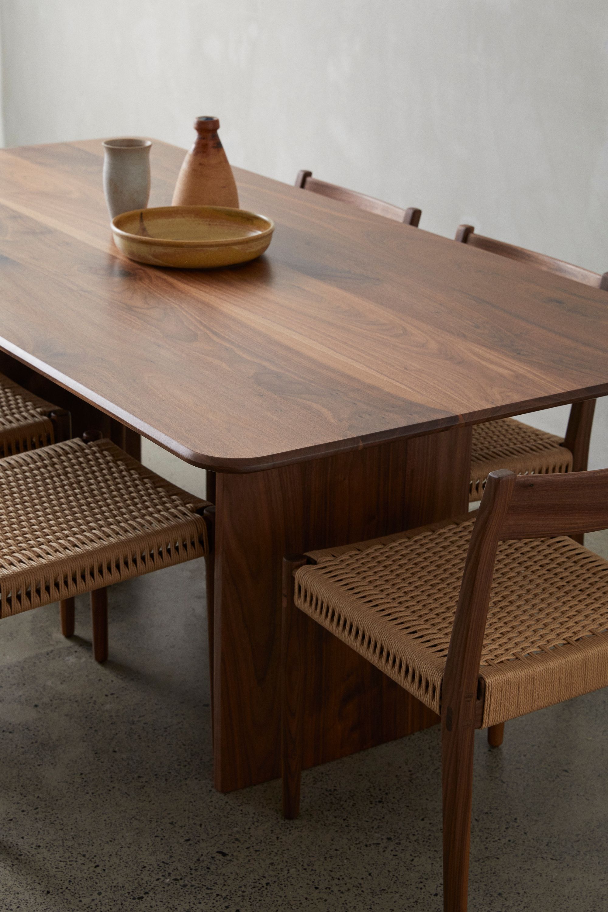 A warm and inviting dining setup showcasing a rich wooden table with a smooth finish. Atop the table are ceramic pieces, including a tall cylindrical vase, a brown bottle-shaped container, and a shallow bowl in an earthy hue. The table is accompanied by chairs with matching wooden frames and woven rattan seats, creating a harmonious blend of textures. The terrazzo-style flooring provides a neutral backdrop, complementing the natural and minimalist aesthetic of the scene. The overall ambiance suggests a serene and rustic elegance.