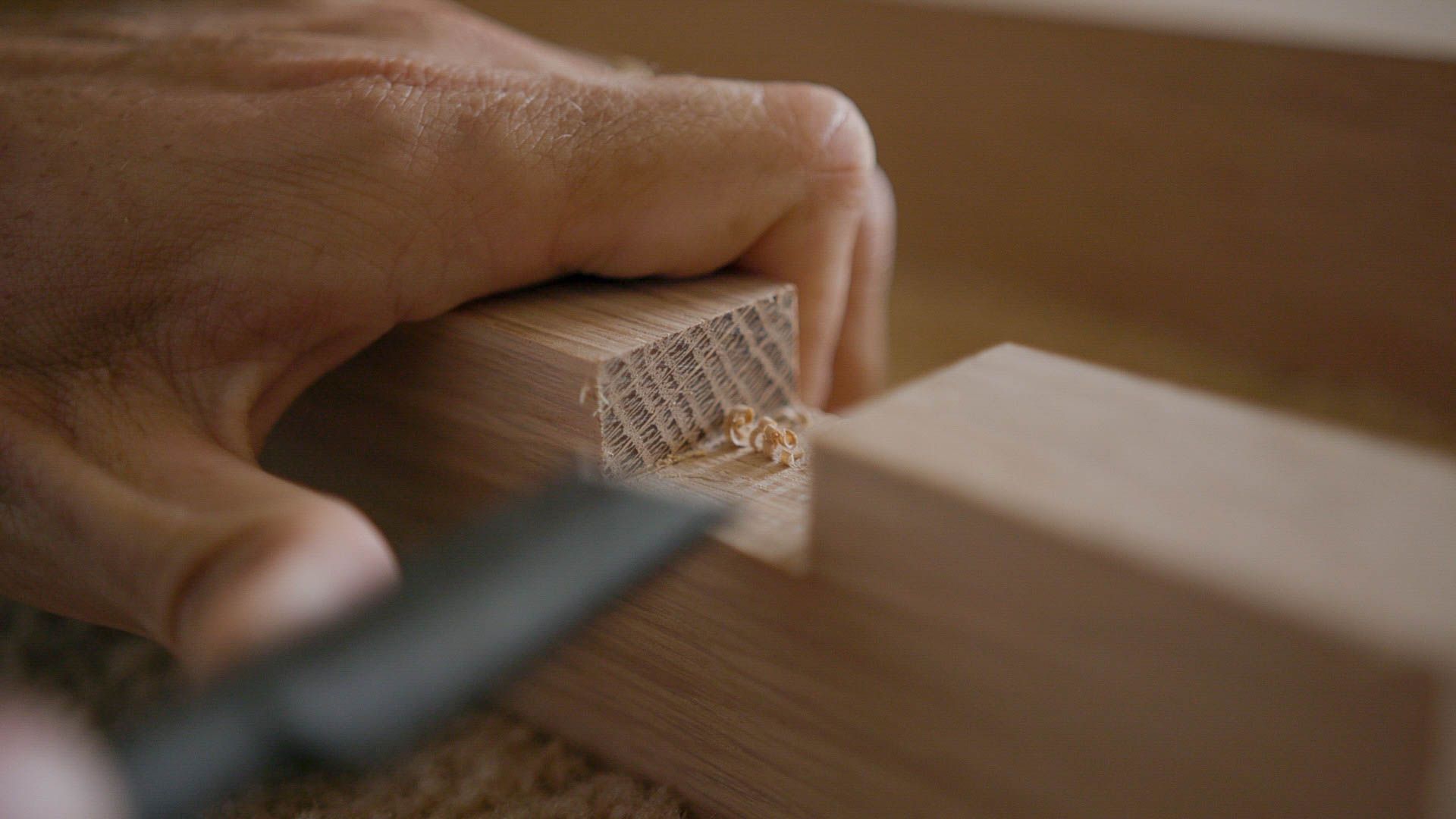A close-up shot of hands skillfully working on a wooden block, using a sharp chisel to carve or shape it. The focused craftsmanship is evident in the fine details of the wood shavings and the precision of the tool. The background is softly blurred, emphasising the main activity.