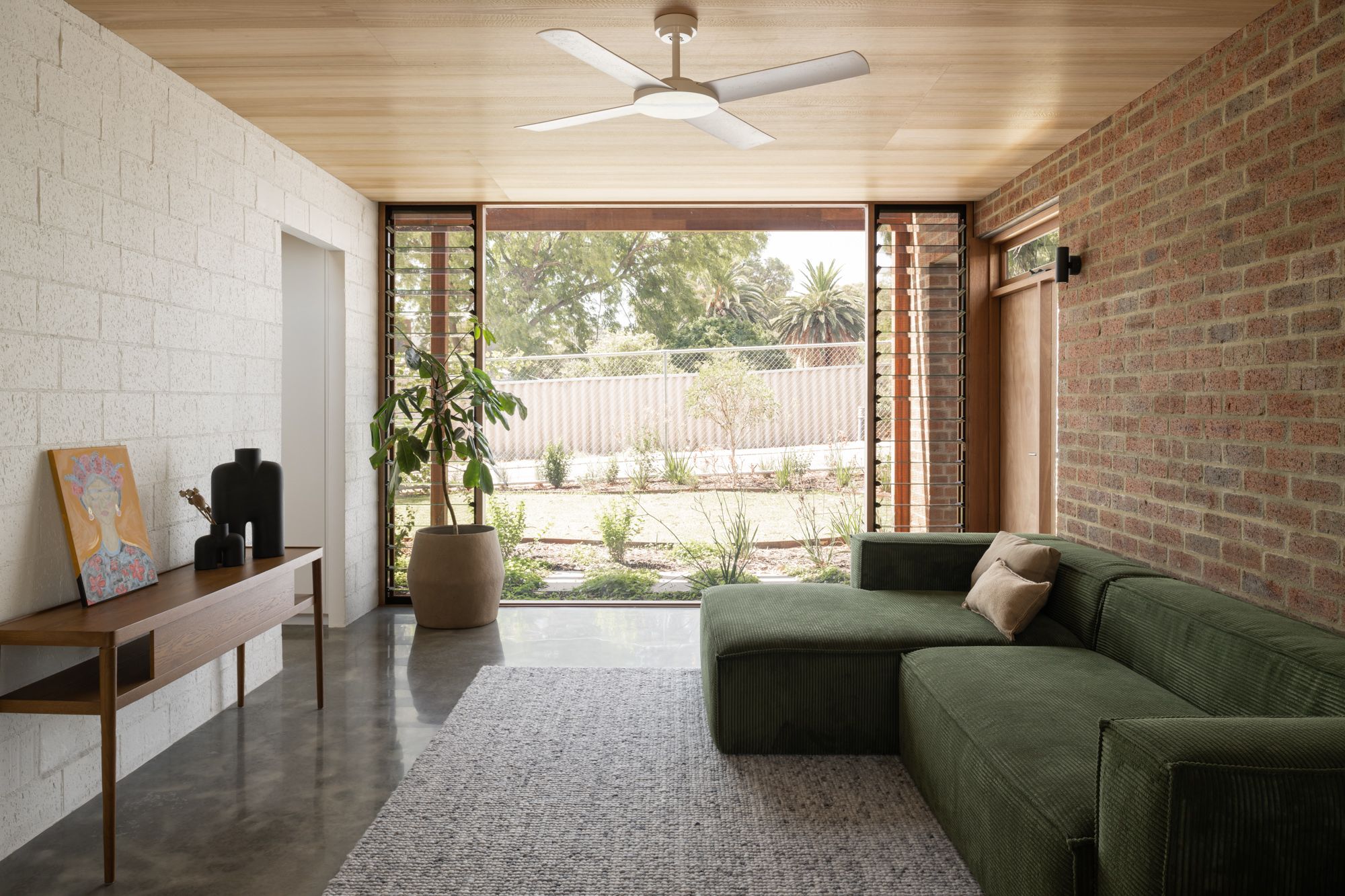 Farrier Lane Home MDC Architects. Brick walled room with polished concrete floors and a timber ceiling. There is a green couch, a timber sideboard and a large potted plant. There is glass plantation shutters opening on to a green backyard. 
