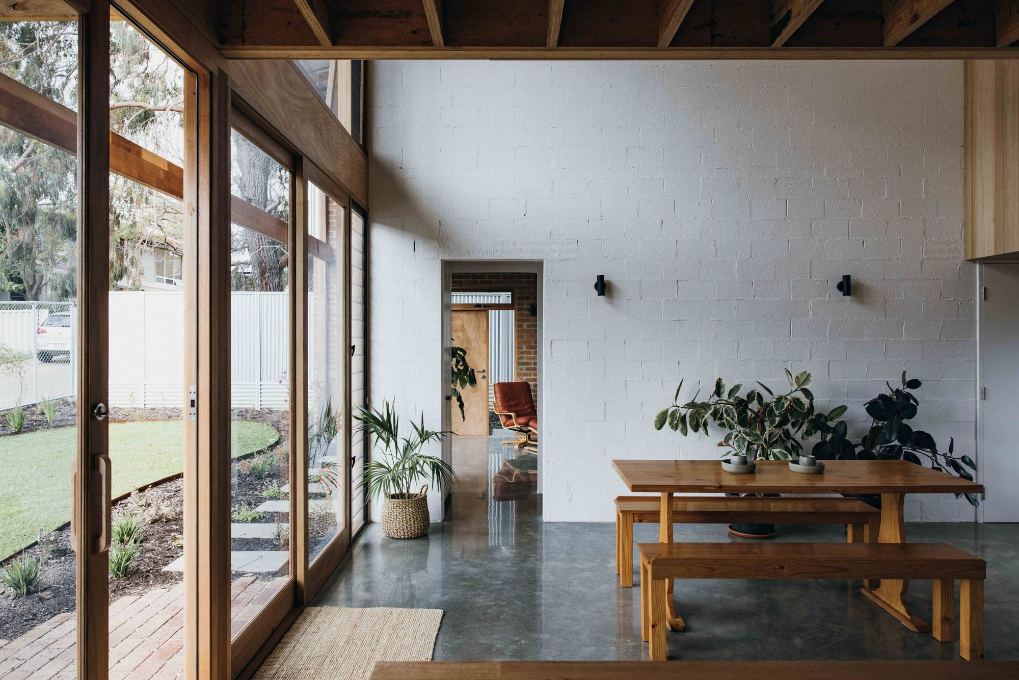 Farrier Lane Home by MDC Architects. Room with polished concrete floors, brick walls painted white and floor-to-ceiling timber windows frames and doors. Polished timber booth seating table in the centre of the room with two rubber plants behind it. Open doorway leading into a living room with a red chair. 