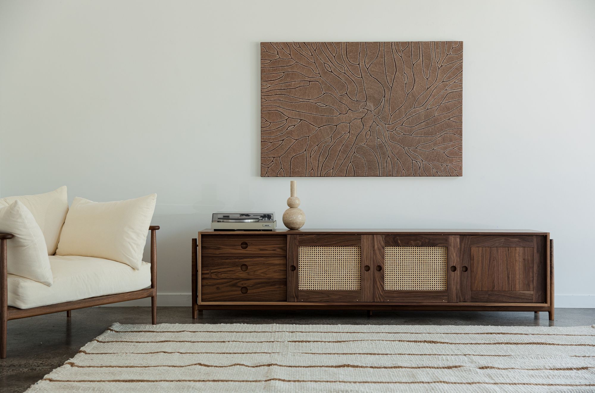 J.D Lee furniture: A minimalist living room setting featuring a light-colored sofa with plush white cushions, positioned adjacent to a finely crafted wooden console with wicker detailing on its doors and a sleek modern turntable on top. Above the console hangs a large square art piece, illustrating intricate abstract wooden patterns. The floor beneath is adorned with a textured off-white rug with wavy patterns, complementing the overall serene ambiance of the room.