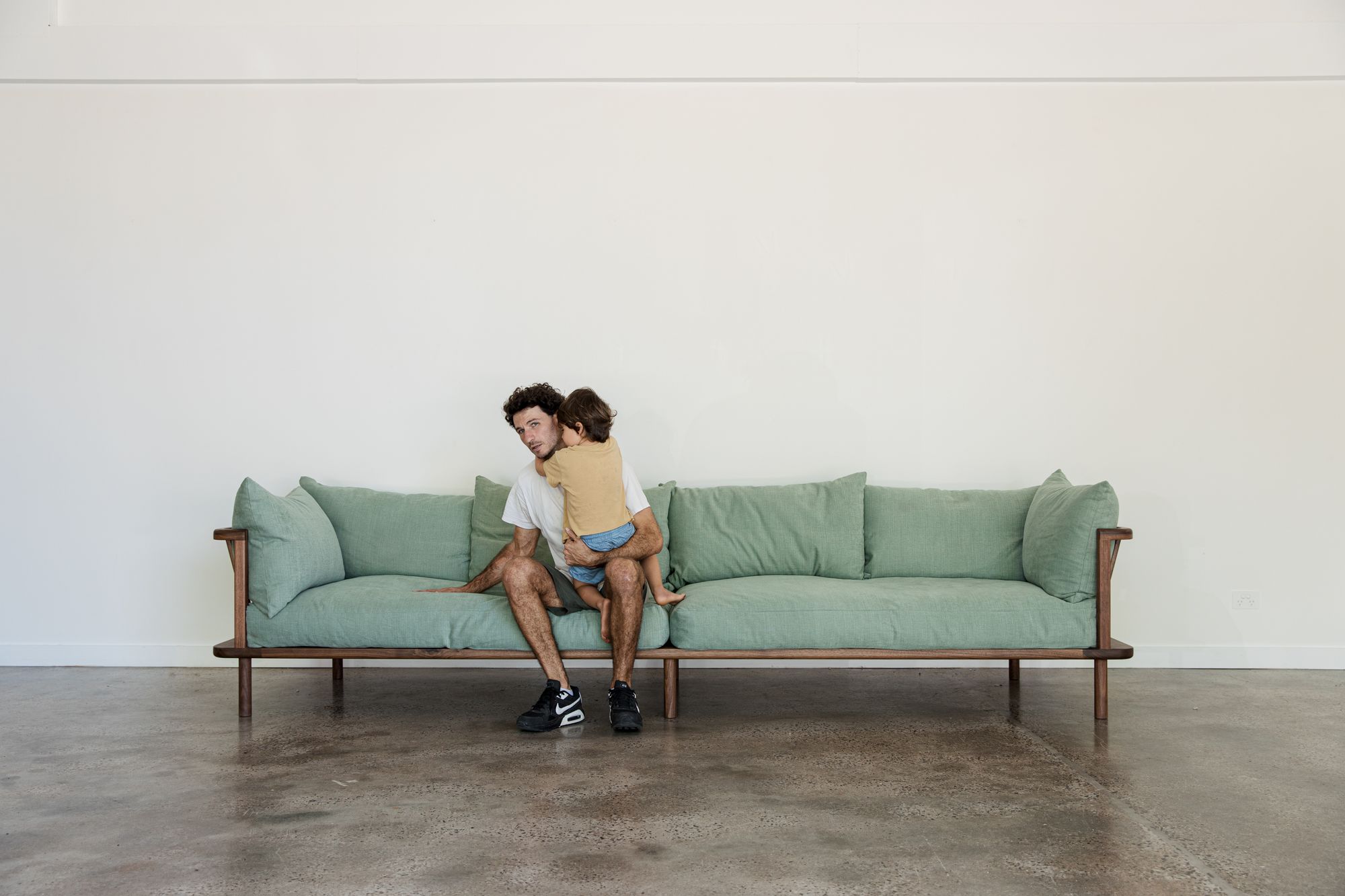  J.D Lee Furniture. A spacious room with a minimalist design, featuring a wooden-framed couch adorned with soft, forest green cushions. Seated on the couch is a jeremy founder of JD.lee  in a white shirt, hugging his  child in a mustard-colored top.  The polished concrete floor reflects natural light, complementing the muted palette of the room and emphasizing the simplicity and coziness of the setting.