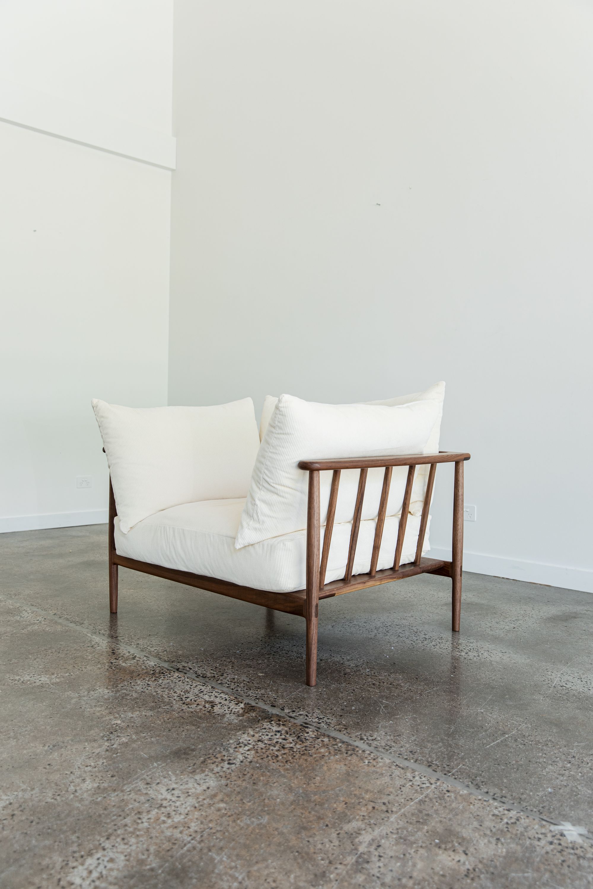 J.D Lee Furniture.A contemporary wooden lounge chair with a minimalist design sits gracefully against a backdrop of white walls. The chair features a deep seat cushioned with soft white pillows, providing a stark contrast to its rich brown frame. Its backrest consists of evenly spaced wooden slats, adding an element of geometric visual interest. 