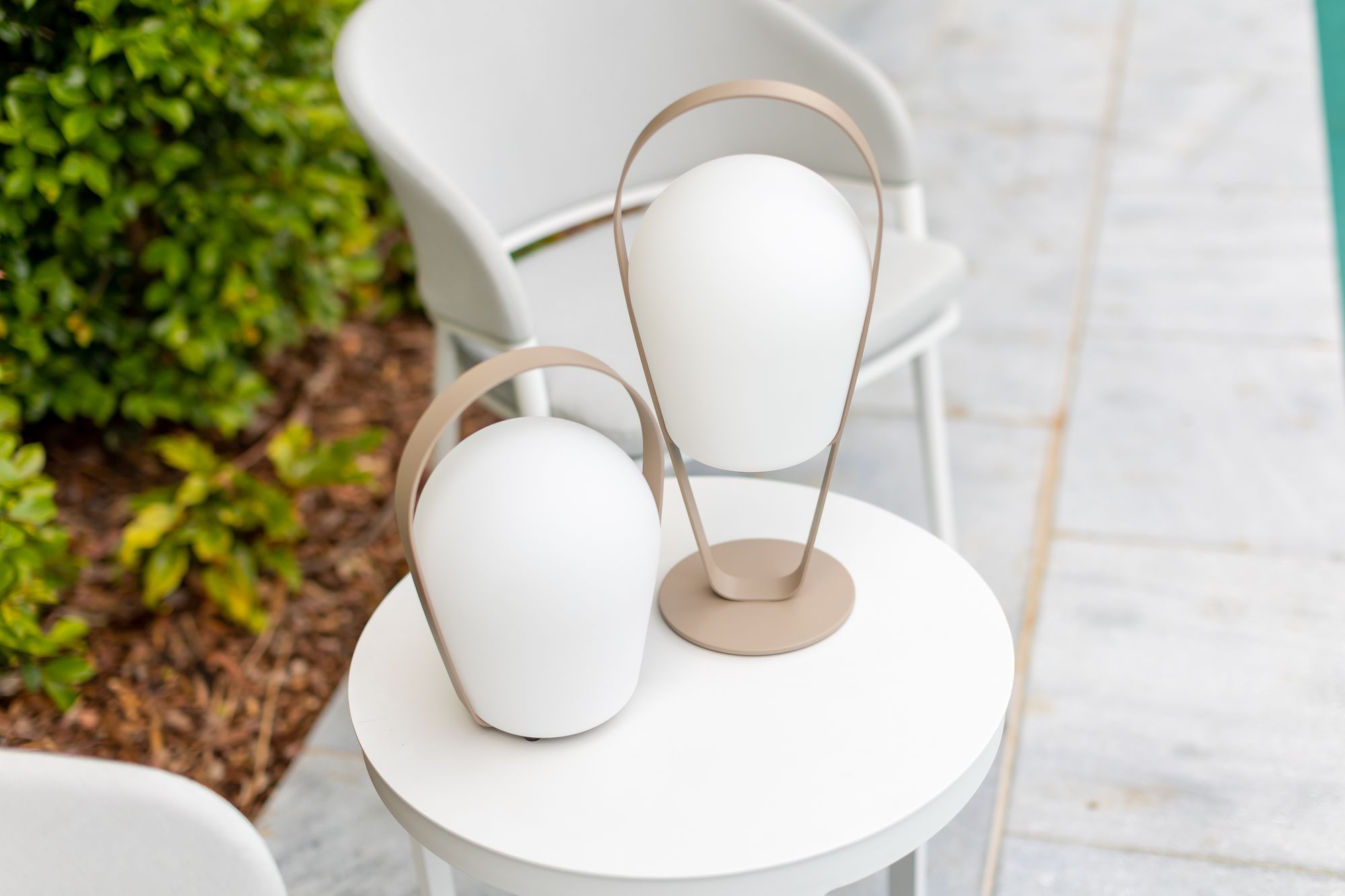 Remarkable Outdoor Living: Bobby Outdoor Aluminum Lamp Low.This image features two modern portable LED lamps placed on a round white table. The lamps have a minimalist design, with matte white diffusers and a sleek bronze-colored handle that curves around the diffuser. The handle serves both as a design element and a functional part, making it easy to carry the lamps. The white and bronze colors suggest a contemporary and elegant aesthetic, fitting well for modern outdoor or indoor living spaces. The greenery in the background provides a nice contrast and indicates that this setting is outdoors, perhaps on a patio or deck.