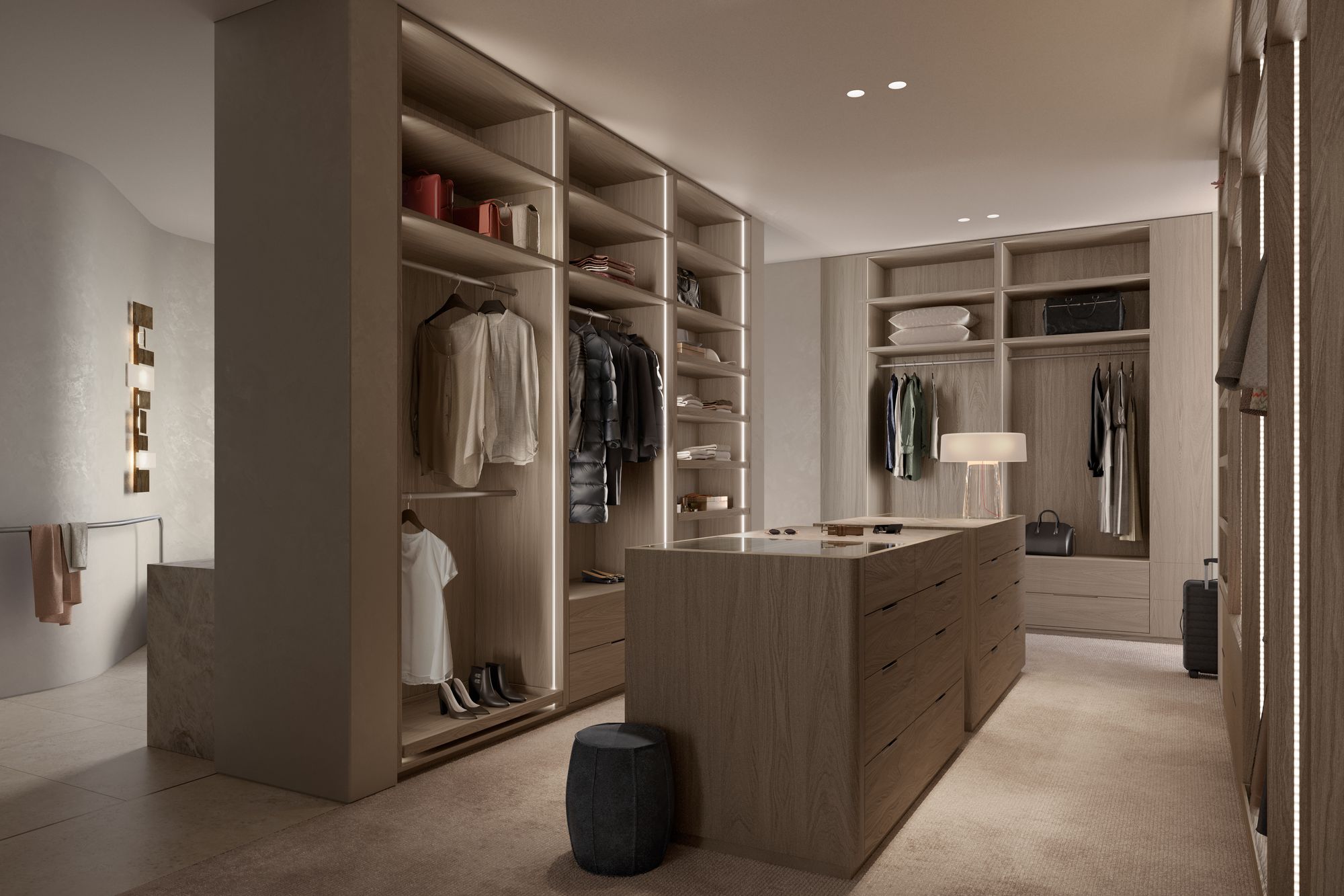 Como Toorak by Prime Edition x Jolson.This image shows a modern, well-organized walk-in closet with natural wood tones and soft lighting, creating a warm and luxurious atmosphere. The space is designed with ample shelving and hanging areas for clothes, an island dresser with drawers for additional storage, and a seated pouf for comfort. Decorative elements like a stylish table lamp, wall art, and a selection of handbags and shoes add a personal touch to the space. The use of mirrors enhances the feeling of spaciousness, and the closet's open design allows for easy access and a display of the wardrobe within.
