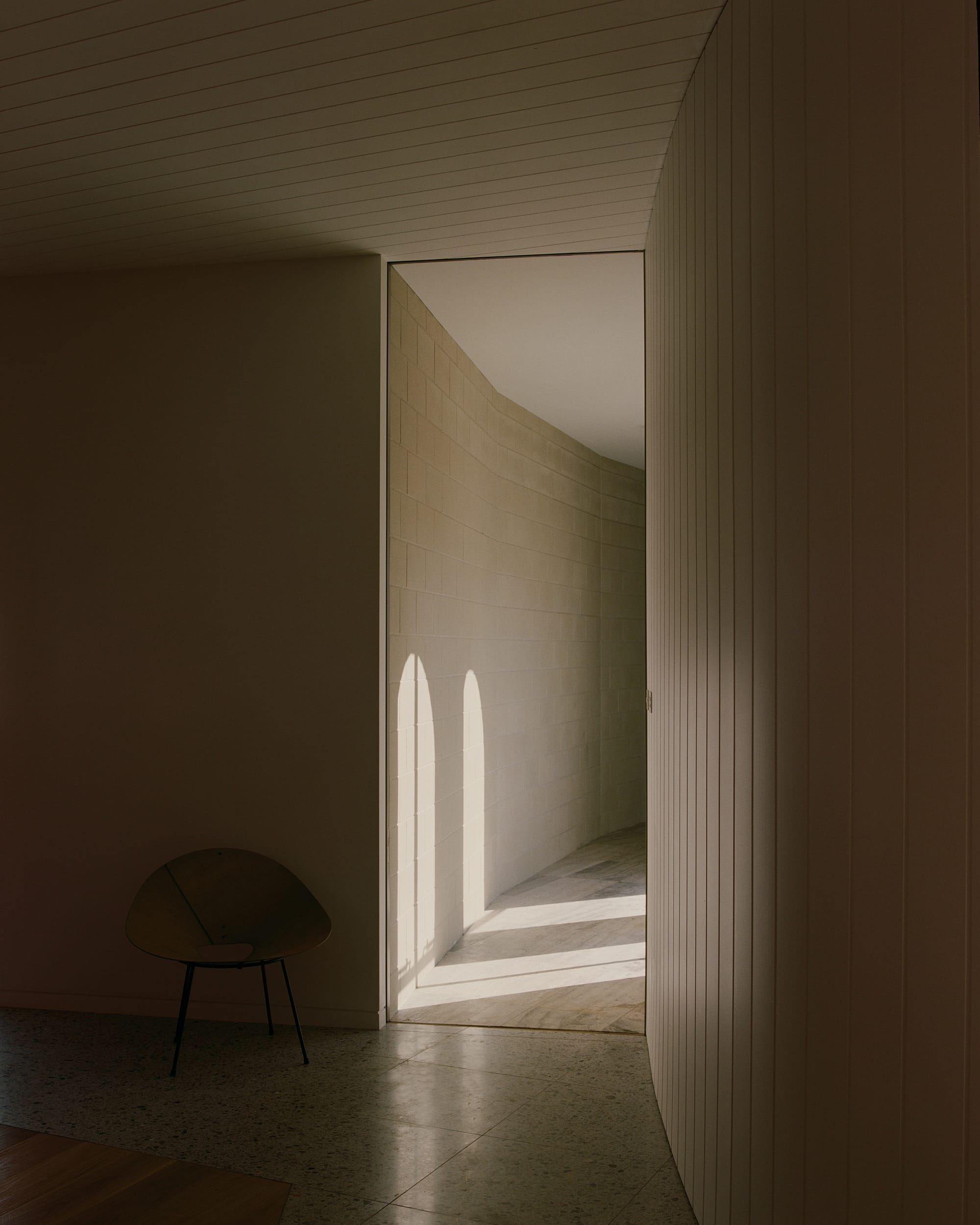 indoor corridor with white walls, featuring shadow patterns created by the sunlight coming through arched openings. A black chair is visible in the corner.