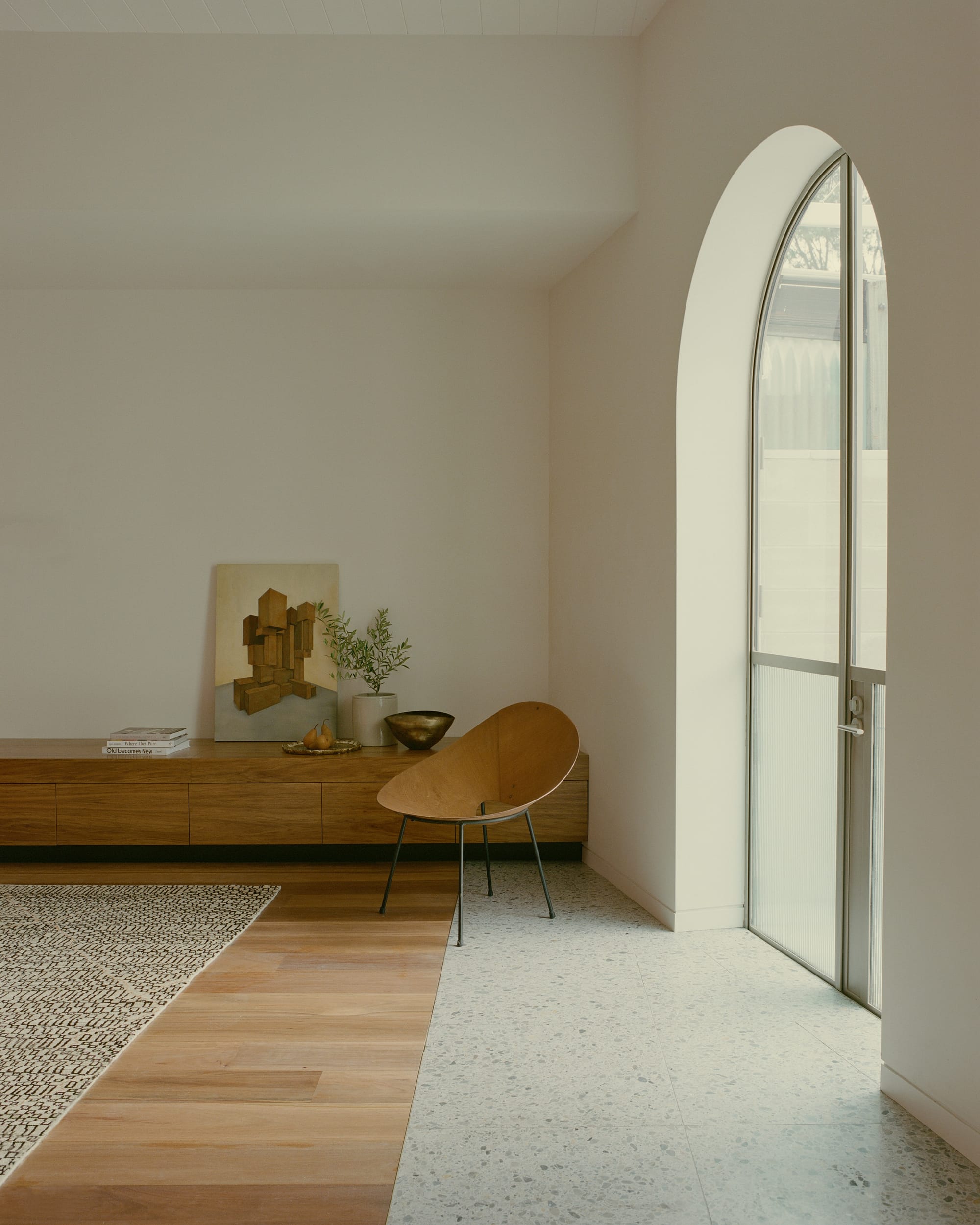  Inside the building, a simple, well-lit room with a large arched window, a wooden console table, a bench, and minimalistic decor is shown.