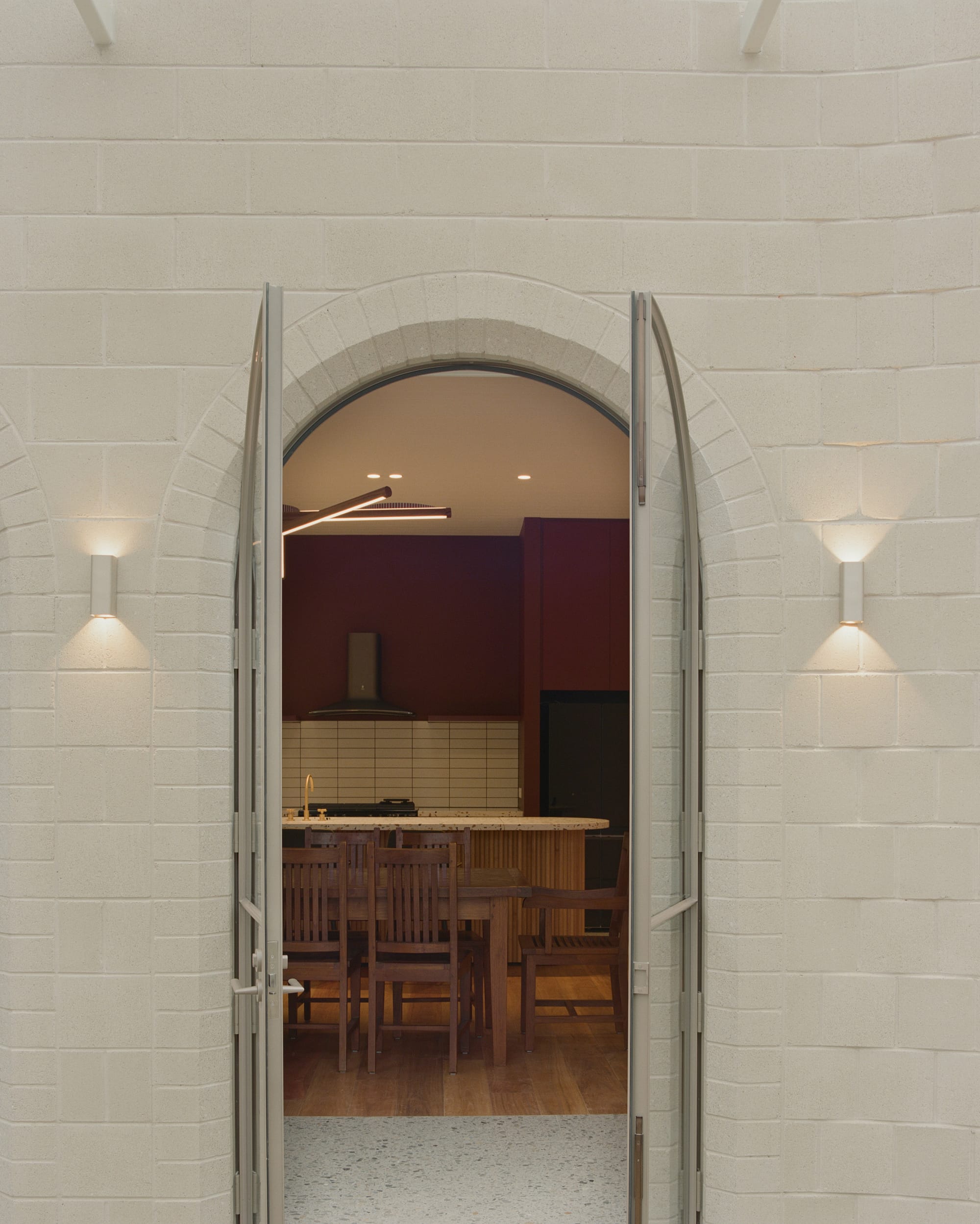 The focus is on an arched double door set in a white brick wall, leading into an interior space with a wooden dining table and chairs. The indoor lighting casts a warm glow contrasting with the natural light outside