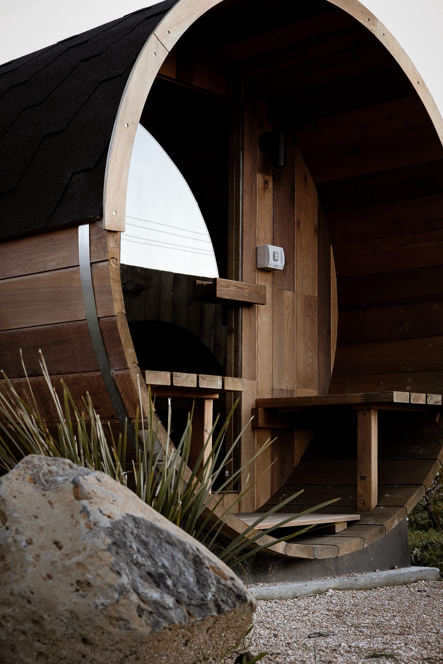 Just Down the Road. Photographer: Studio Winslow.Close-up view of the entrance to a barrel-shaped wooden sauna with an arched opening, revealing the interior benches and wooden structures, positioned on a gravel surface with a large rock in the foreground.