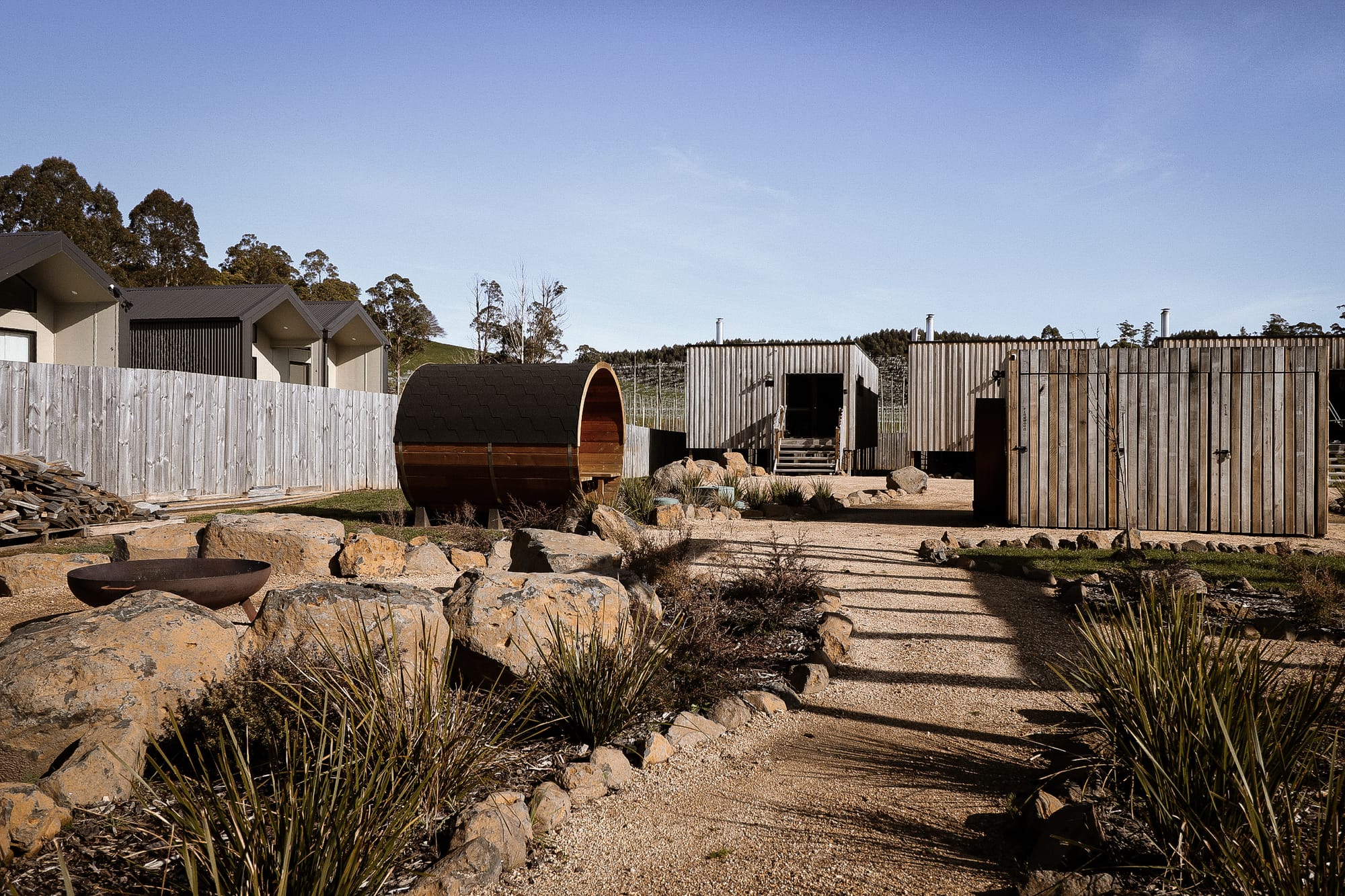 Just Down the Road. Photographer: Studio Winslow. A rural landscape with a wooden fence and a large, cylindrical wooden sauna with a curved roof, set amongst an arid garden with sparse vegetation under a clear blue sky.