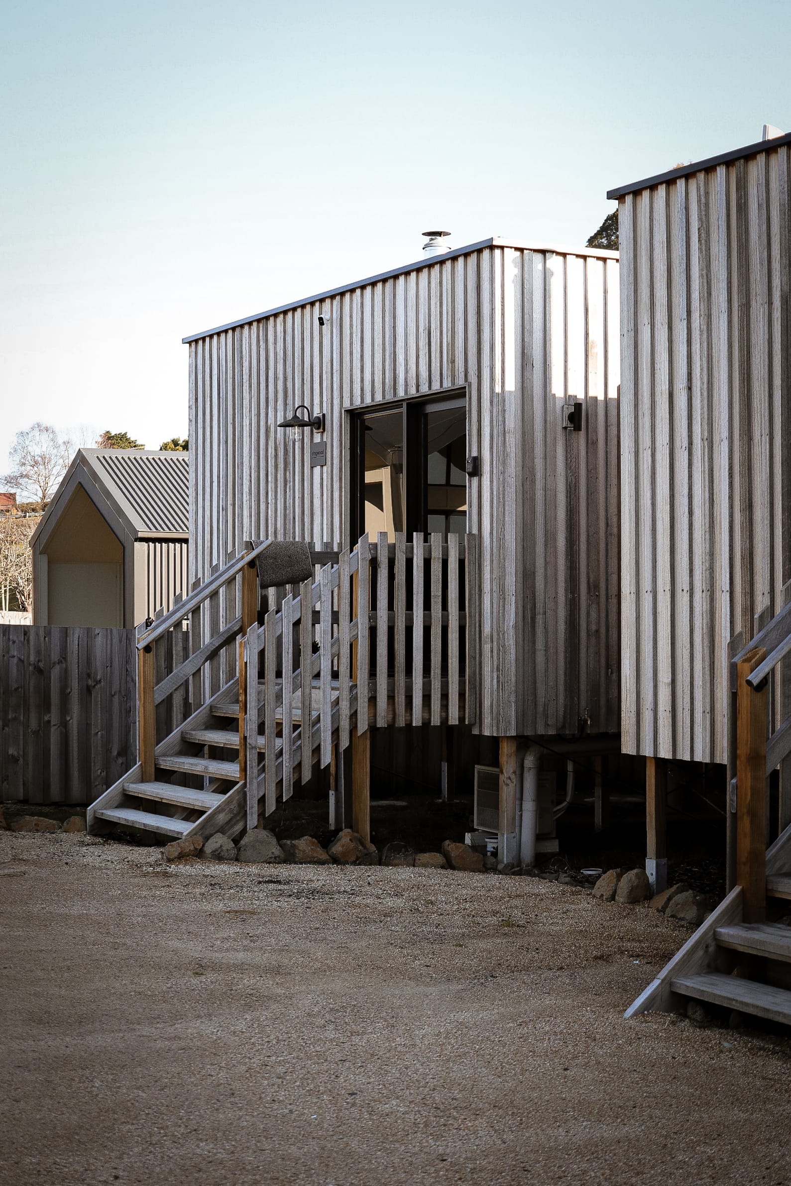 Just Down the Road. Photographer: Studio Winslow.  A modern wooden cabin with corrugated metal siding, featuring a stairway leading up to a deck with a door, set in a residential area with other houses nearby and a clear blue sky above.