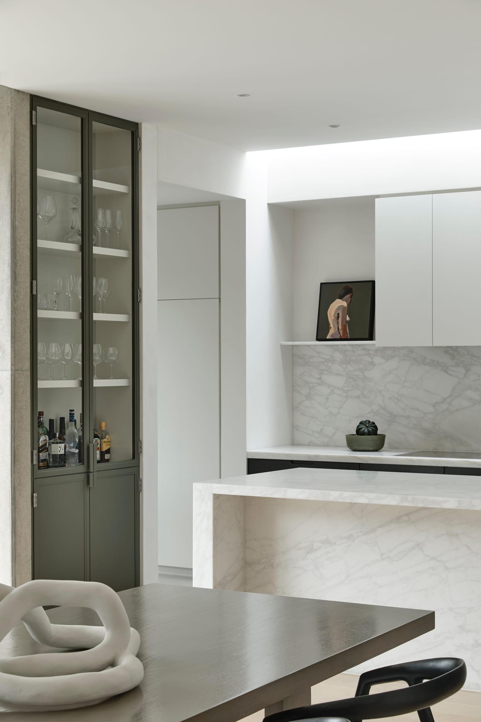 RE Residence by Inglis Architects.minimalist kitchen space with a marble countertop extending to a backsplash. On the left, a tall cabinet with glass doors reveals an organized display of glassware and bottles, suggesting a built-in bar area. To the right, a niche in the wall displays a small sculpture, adding a personal touch. A chunky, sculptural grey knot decorates the foreground, sitting on a wooden table accompanied by a sleek black chair, blending functional design with artistic elements. The overall palette is neutral, with natural light softening the space.