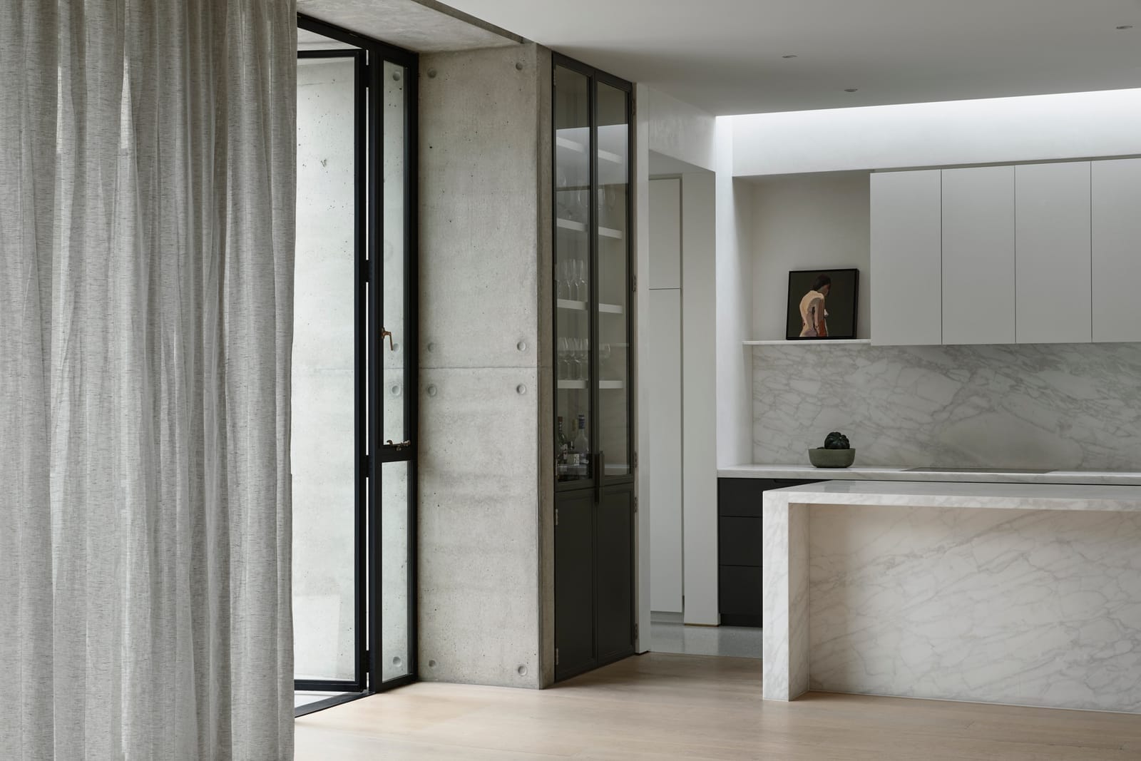 RE Residence by Inglis Architects.partial view of a living room leading to a kitchen. Sheer curtains frame sliding glass doors that reveal the edge of a kitchen island with a marble countertop. A small sculpture is placed on a shelf, and white cabinetry is visible in the background.