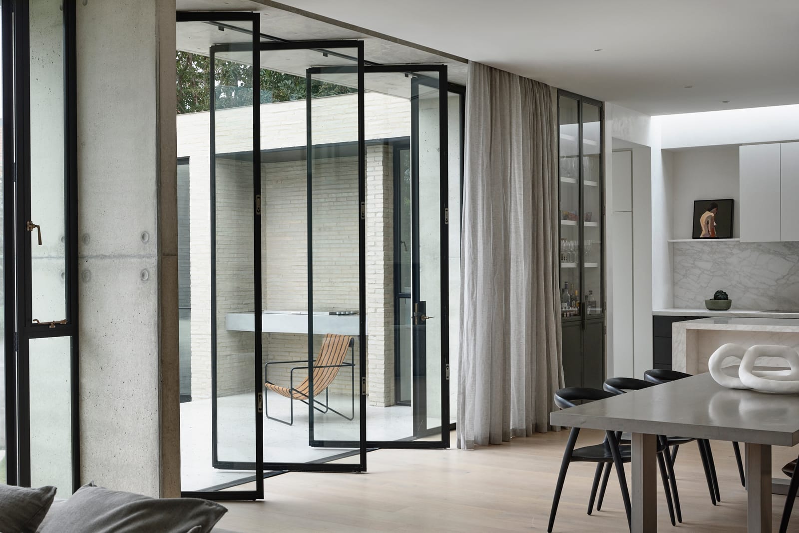 RE Residence by Inglis Architects.iew from the inside looking out through black-framed glass doors onto a patio. The doors are ajar, allowing a glimpse of a comfortable chair on the patio. Inside, the focus is on a dining area with a dark wooden table and black chairs, complemented by the same sheer curtains as the top view, tying the two spaces together visually.
