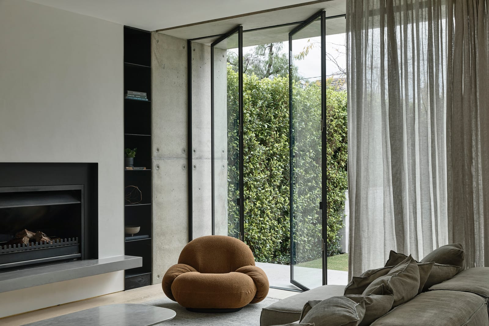 RE Residence by Inglis Architects cozy corner of a modern living room with large glass sliding doors that open to an outdoor area with greenery. Sheer curtains filter the natural light, enhancing the tranquil atmosphere. A contemporary, round, brown chair sits in the foreground, and a plush, grey sofa adorned with cushions is partially visible. A minimalist fireplace anchors the space to the left, adding warmth to the setting.