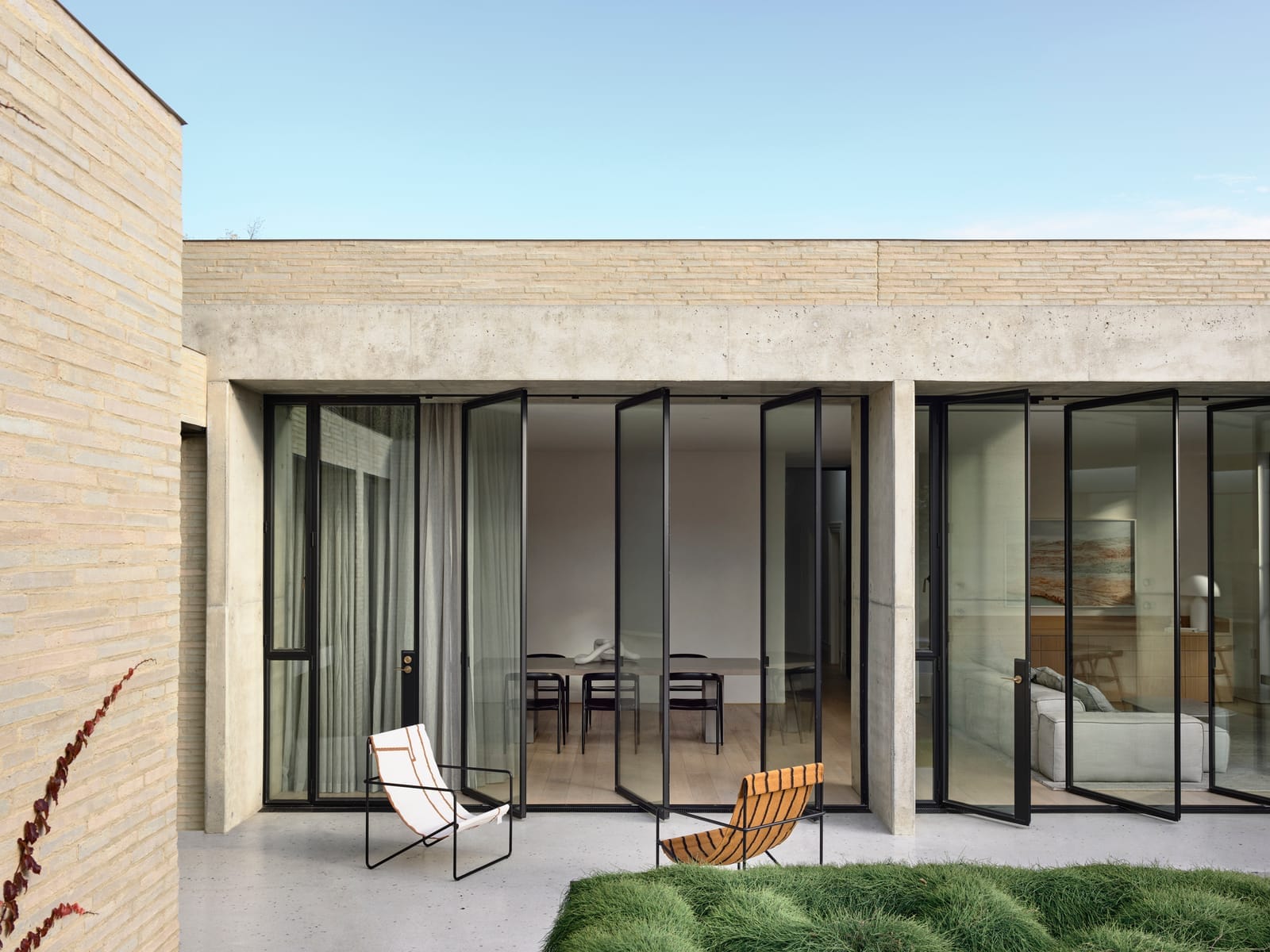 RE Residence by Inglis Architects.  modern residence with a clear sky above. The facade is minimalist with large glass doors and windows framed by stone walls. A single chair and a woven lounge chair are placed on the concrete patio.