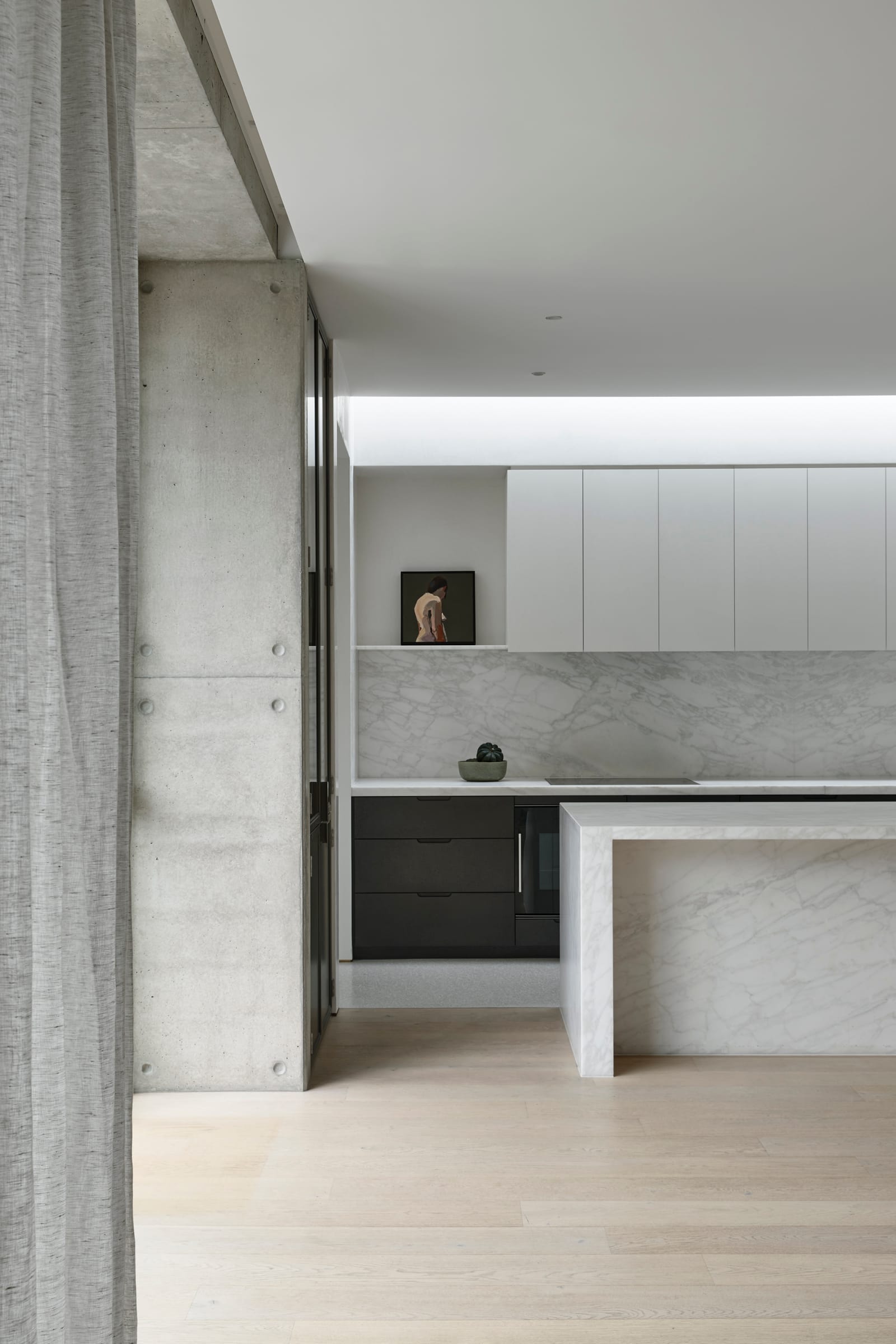 RE Residence by Inglis Architects.interior view of a modern kitchen with minimalist design. A marble island with a waterfall edge is the centerpiece, matched by a marble backsplash. The room is framed by light wooden flooring and a concrete ceiling, with white cabinetry on the wall and a black cupboard below the counter. A small sculpture is visible on a shelf, adding a personal touch to the space.