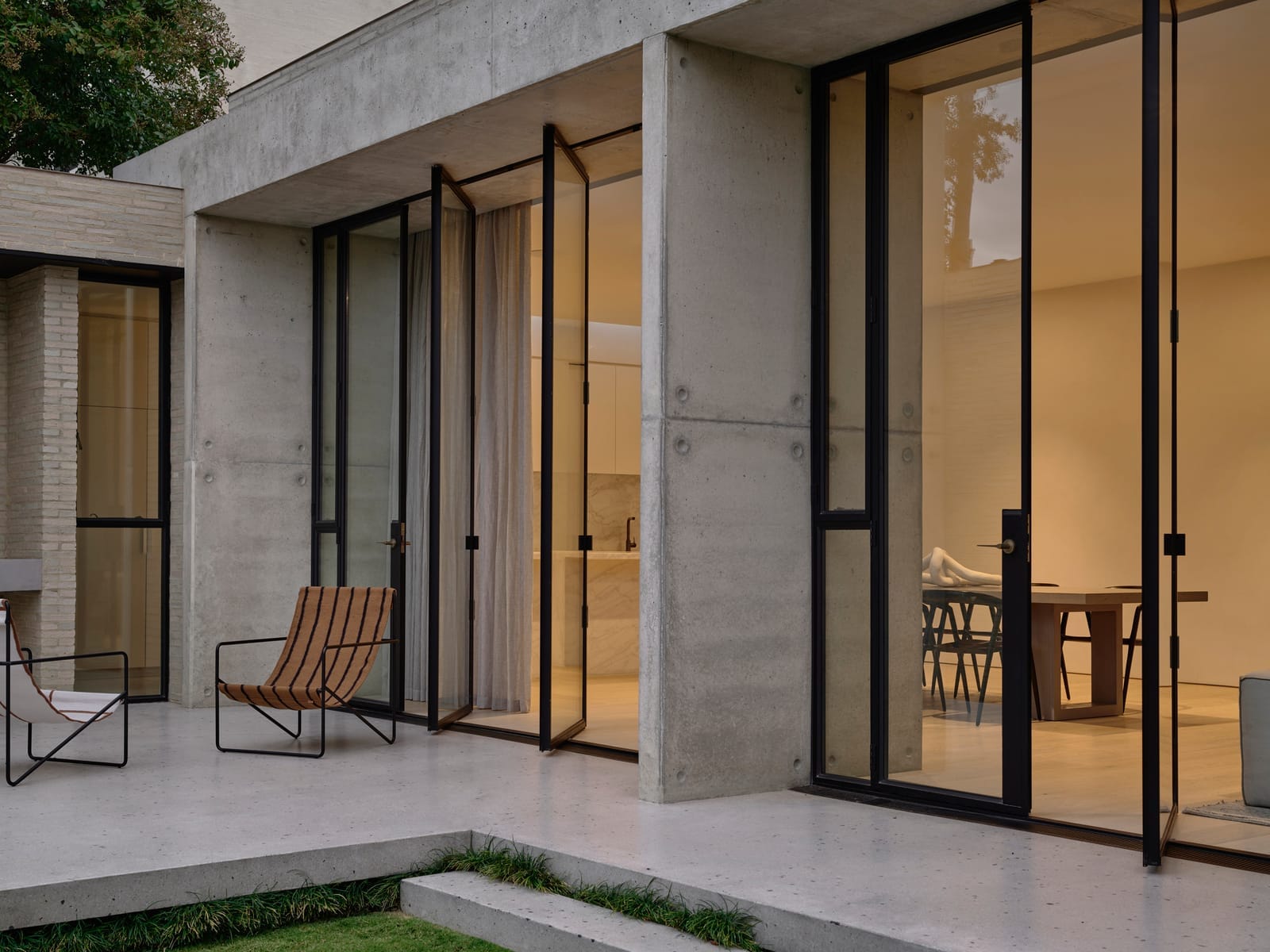 RE Residence by Inglis Architects.The image depicts an evening view of a modern architectural residence with a concrete facade. Large floor-to-ceiling glass doors with black frames open onto a patio. One of the doors is ajar, inviting a look into the warmly lit interior. On the patio, there is a metal frame chair with a wooden slat seat and backrest, enhancing the minimalist aesthetic of the home.