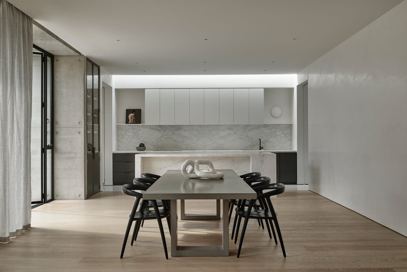RE Residence by Inglis Architects.The first image shows a spacious and modern dining area within a minimalist kitchen setting. A simple wooden dining table is paired with black chairs, situated on light wooden flooring. White cabinetry and a kitchen counter with a marble finish define the kitchen space in the background, with a glimpse of a hallway to the left, featuring a concrete wall.