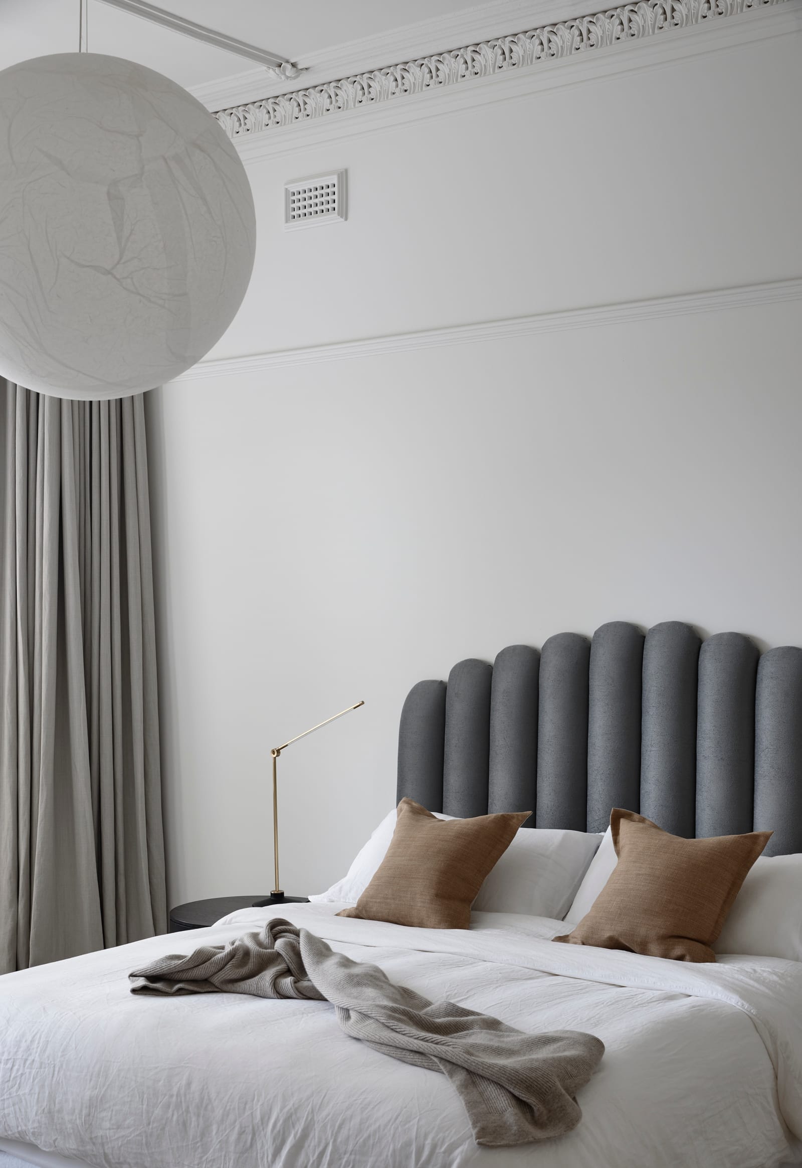 RE Residence by Inglis Architects.The room has a high ceiling and is painted in a soft white, with a large globe pendant light hanging off-center. The bed features a unique, cylindrical grey headboard, and is dressed with crisp white linens, a grey throw, and two earth-toned pillows. Elegant, floor-length curtains add a touch of luxury to the room. A brass reading lamp on the right completes the setting.