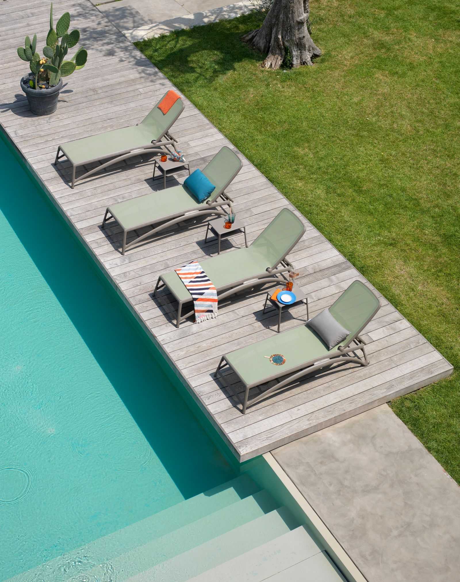 Remarkable Outdoor Living: Nadi Altantico Outdoor Resin Sunlounger. An outdoor pool area is furnished with sleek sun loungers arranged neatly on a wooden deck, complemented by a potted cactus and soft-colored towels, all inviting relaxation under the open sky.