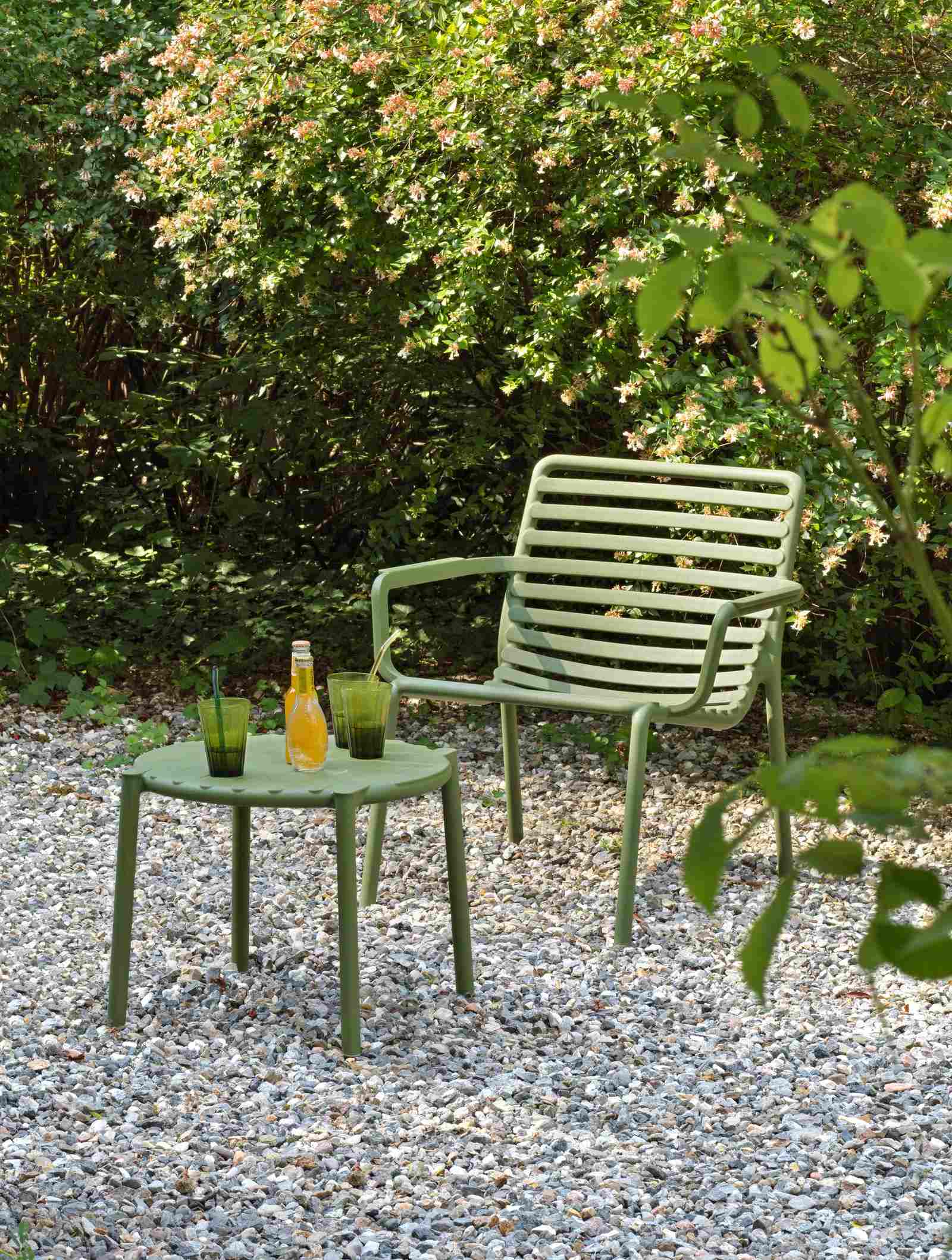 Remarkable Outdoor Living: Nadi Doga Outdoor Resin Balcony Relax Chair.This image shows a calming garden scene with a single sage green chair and a matching round table. The furniture is set on a gravel surface, which gives it a rustic and natural look. On the table, there are two tall glasses, likely containing a refreshing green beverage, and a small bottle, which could be a syrup or juice to mix with the drinks. The lush greenery in the background, consisting of various shrubs and plants, indicates that this outdoor space is designed for relaxation, perhaps as a quiet corner in a garden where one could enjoy a drink and the tranquility of nature.