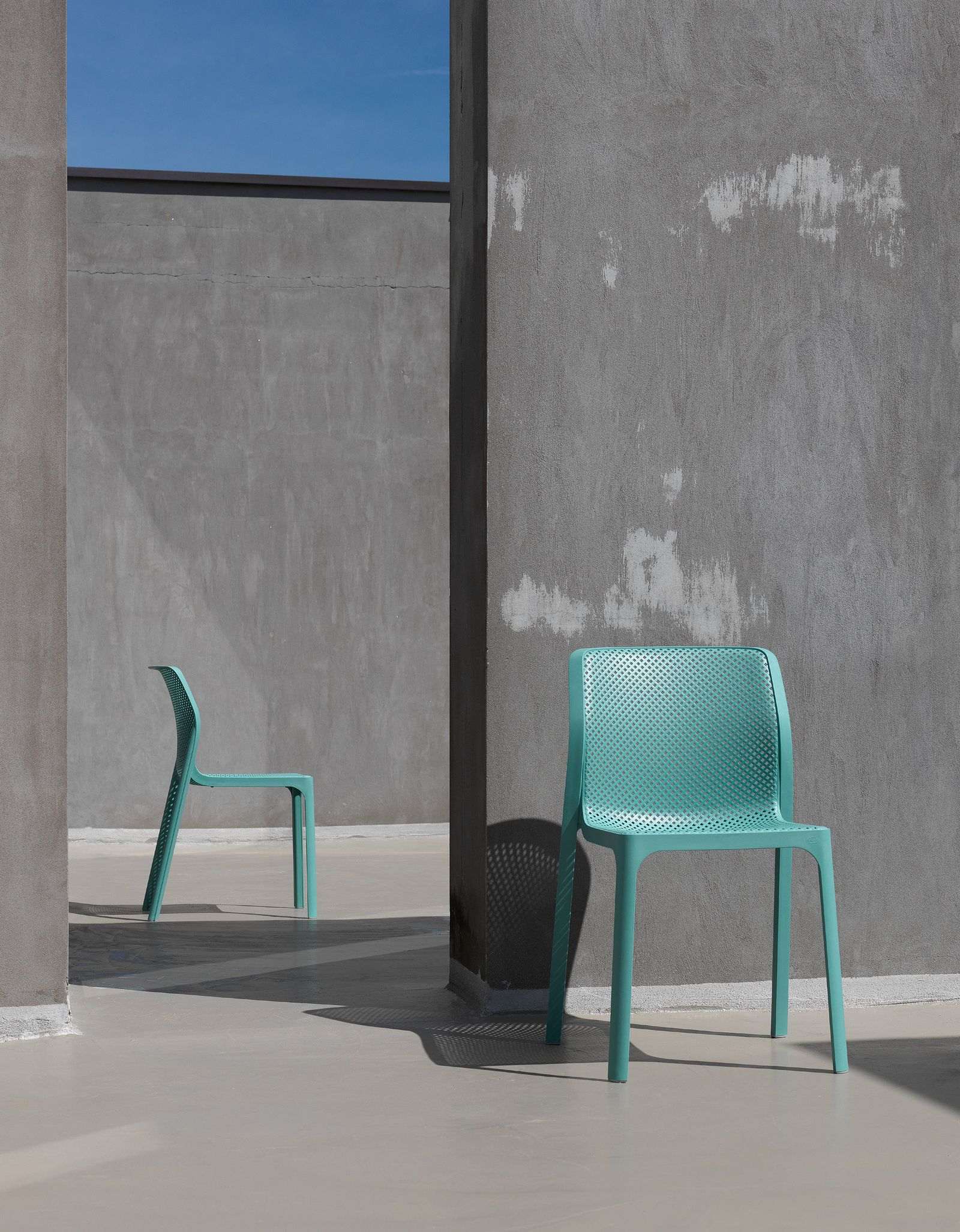 Remarkable Outdoor Living: Nardi Bit Outdoor Resin Dining Chair. Two modern turquoise chairs are positioned on a smooth concrete surface, creating a stark contrast against the gray backdrop, with the clear blue sky peeking from above.