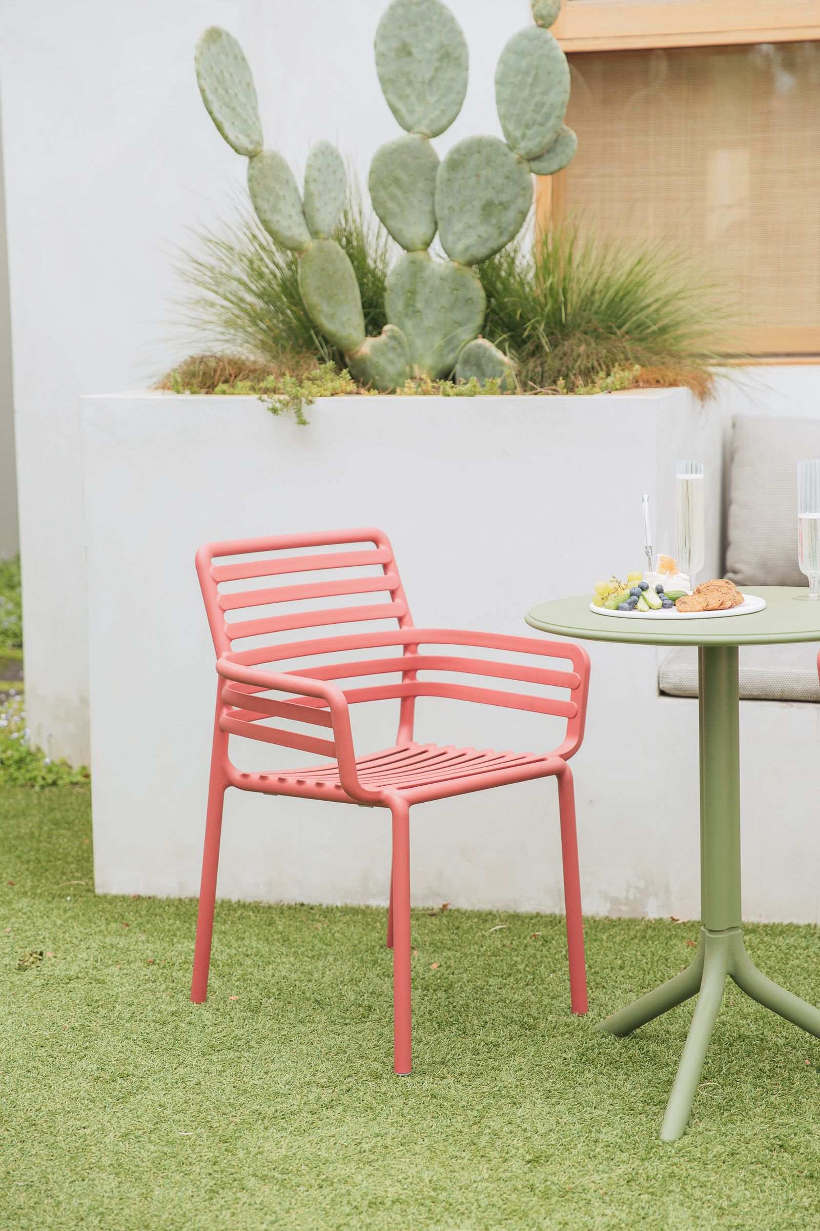 Remarkable Outdoor Living: Nadi Doga Outdoor Resin Dining Armchair. The image presents a neatly composed outdoor setting featuring a coral pink chair with a simple, modern design. The chair is accompanied by a round table with an olive green stand, upon which rests a small plate of cookies, a bunch of grapes, and two elegant champagne glasses, suggesting a prepared snack or light refreshment. In the background, a large cactus within a white raised planter adds a touch of natural greenery to the scene, which contrasts nicely with the chair and table colors.