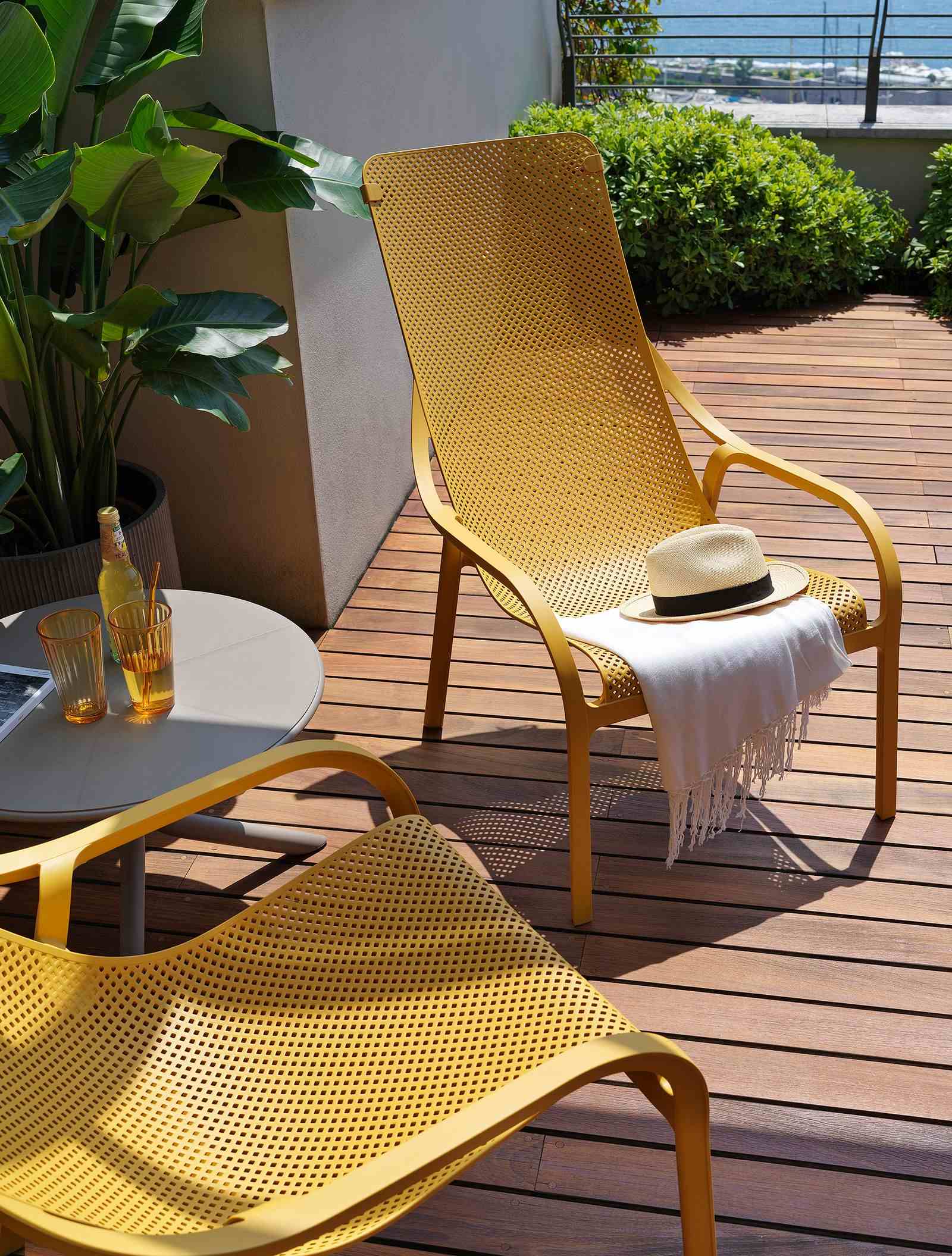 Remarkable Outdoor Living. Nardi Net Outdoor Resin Balcony Lounge Chair. This image shows a vibrant outdoor setting with a yellow perforated metal lounge chair and matching table set against a backdrop of lush greenery and a clear sky. A white hat with a black band rests on the chair, adding a leisurely touch to the scene. The ensemble is situated on a wooden deck, enhancing the warm and inviting ambiance perfect for relaxation or socializing outdoors.