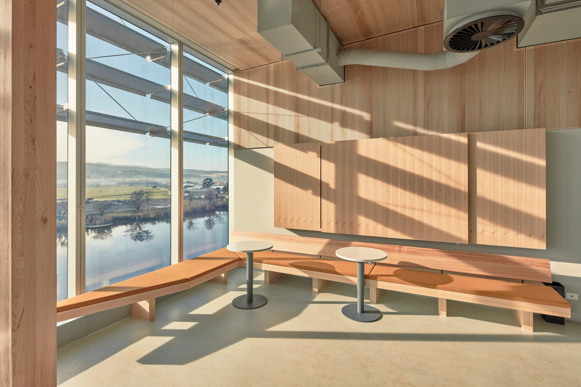 Tasmanian Timber: River's Edge. Photography: Dave Groves. A cozy nook with a large window offering a panoramic view of a serene water body and distant landscape. The room has a minimalist design with two long, wooden benches and a table. The interior is bathed in warm sunlight, casting long shadows on the wooden floor, while the peaceful exterior view suggests a harmonious blend with nature.  