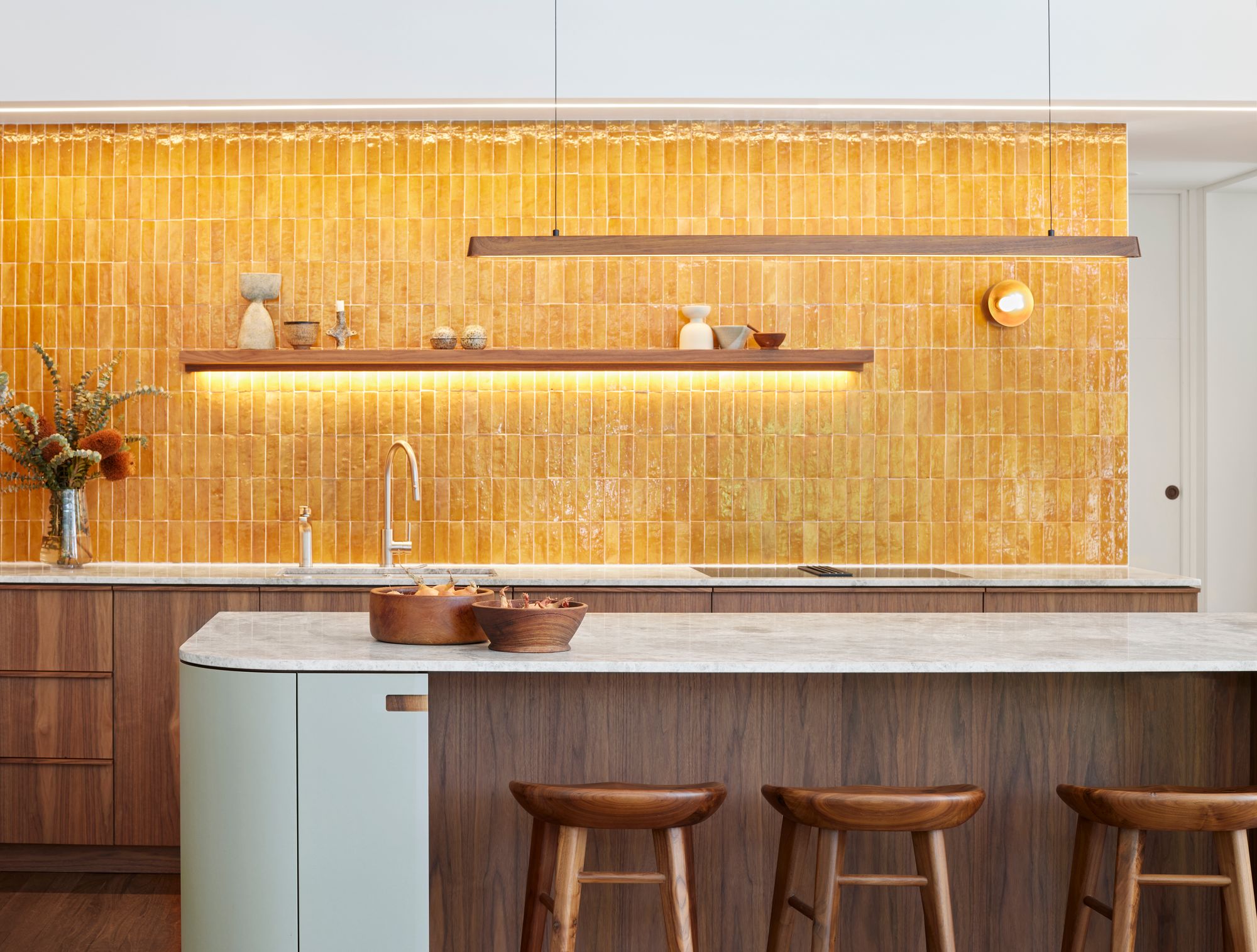  Fluxwood Tenn Pendant. Photography by Fiona Galbraith. The image features a modern kitchen interior with a minimalist design. The focal point is a textured, honey-yellow tiled backsplash that extends from the countertop to the ceiling. Below it, there is a sleek kitchen counter with a marble surface, featuring an undermount sink and a modern faucet. Above the backsplash, a wooden shelf holds various decorative items, and there's a warm, under-shelf lighting that adds ambiance to the space. Three wooden bar stools are placed in front of the kitchen island, which has a base with a pale green finish that contrasts with the rich wooden tone of the cabinetry. The overall aesthetic is clean, warm, and inviting with a touch of rustic charm through the use of natural materials.