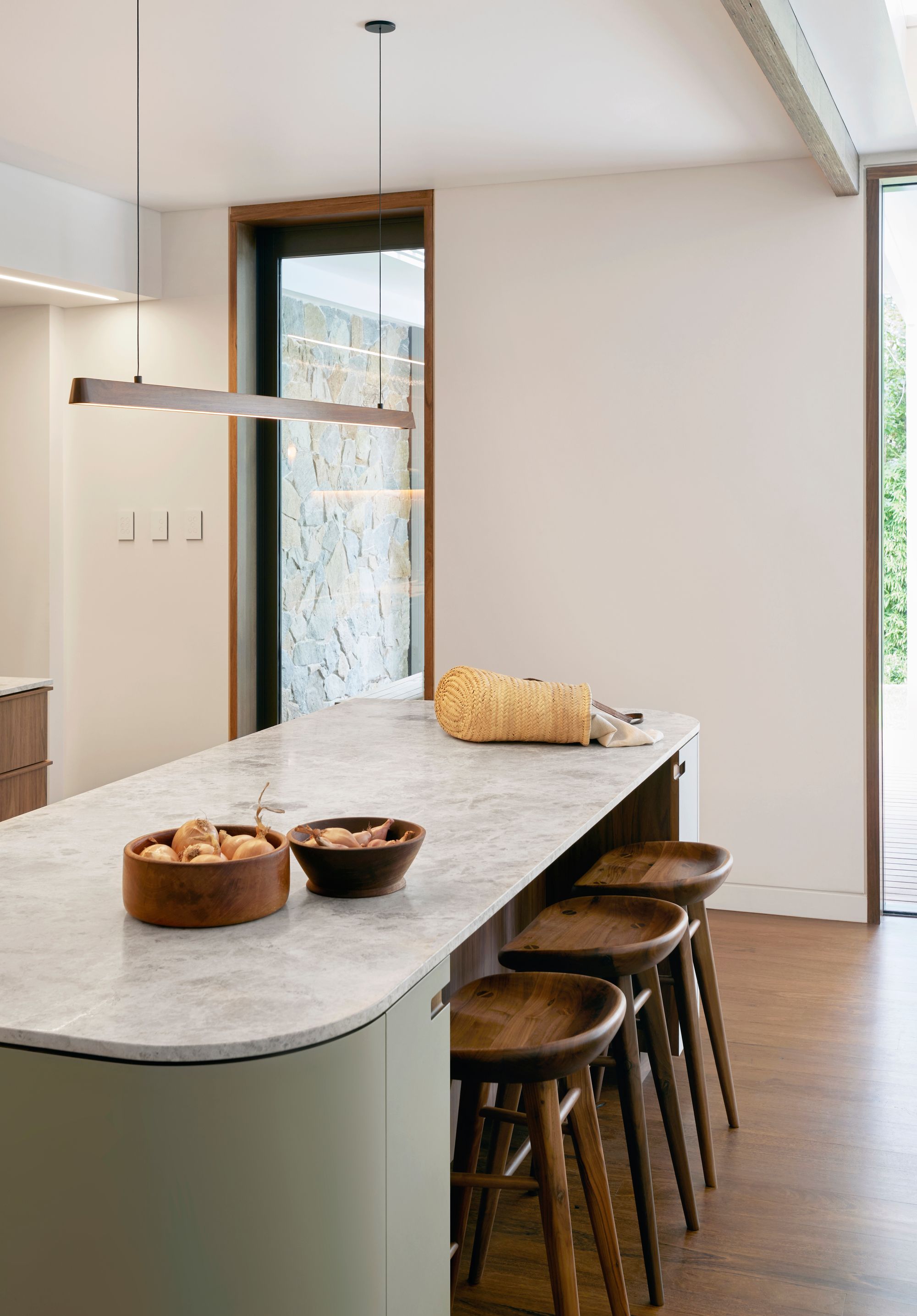  Fluxwood Tenn Pendant. Photography by Fiona Galbraith. The image features a modern kitchen interior with a minimalist design. The focal point is a textured, honey-yellow tiled backsplash that extends from the countertop to the ceiling. Below it, there is a sleek kitchen counter with a marble surface, featuring an undermount sink and a modern faucet. Above the backsplash, a wooden shelf holds various decorative items, and there's a warm, under-shelf lighting that adds ambiance to the space. Three wooden bar stools are placed in front of the kitchen island, which has a base with a pale green finish that contrasts with the rich wooden tone of the cabinetry. The overall aesthetic is clean, warm, and inviting with a touch of rustic charm through the use of natural materials.