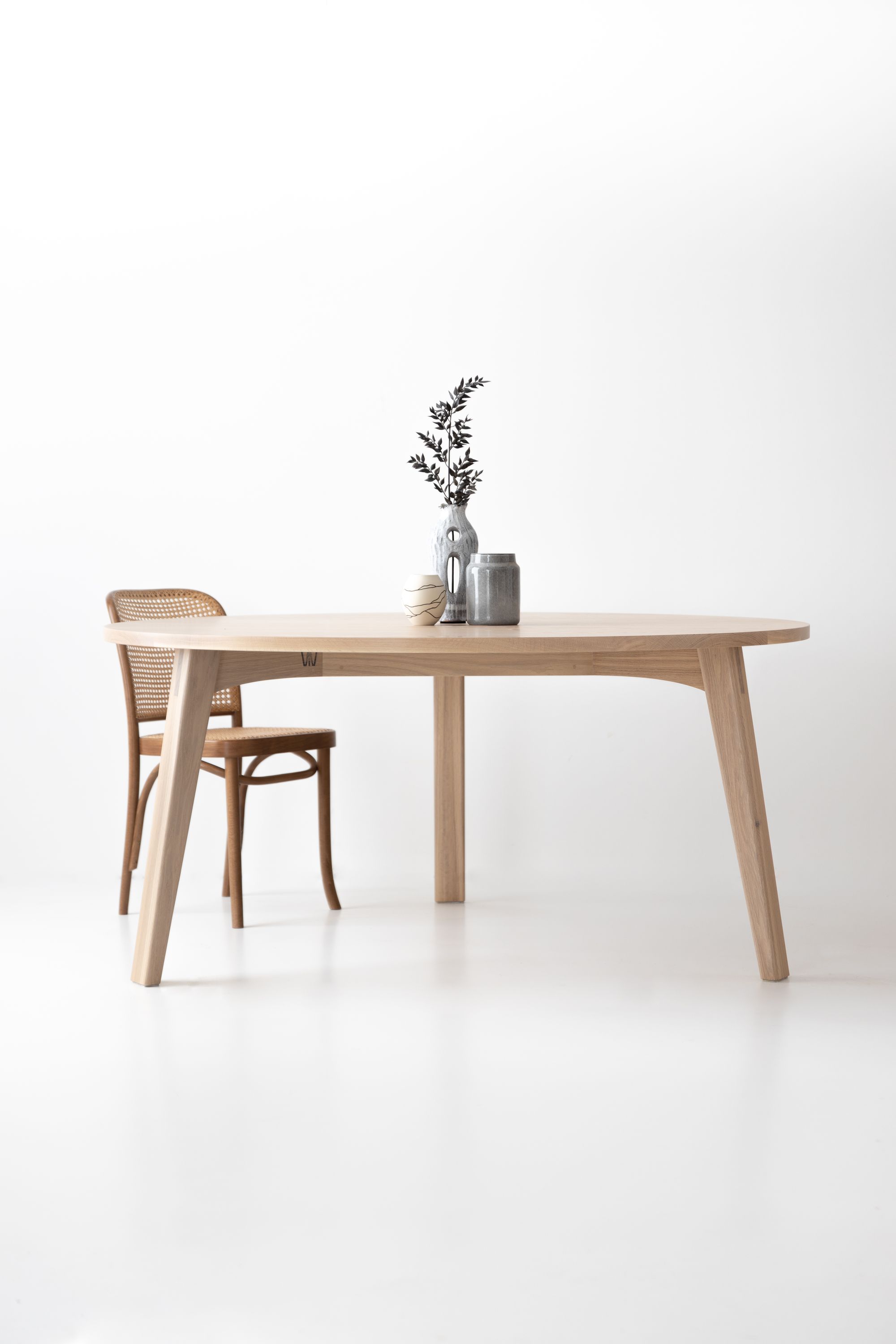 The table is adorned with a small vase holding a single branch with small leaves, alongside a grey pottery piece and a small cup with an abstract design. The white background provides a stark contrast that emphasizes the simplicity and clean lines of the furniture.