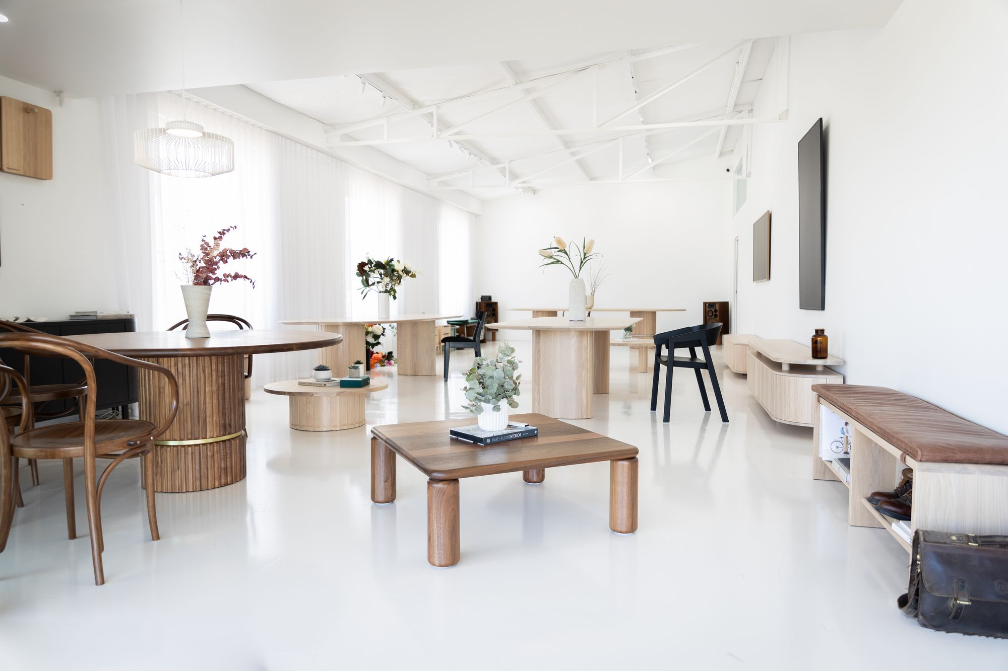 interior showcases a variety of furniture pieces in a white-walled room with a glossy white floor, reflecting the natural light entering through the sheer curtains. The space feels like a curated gallery with a mix of round and rectangular wooden tables, bentwood chairs, and unique bench seating with built-in storage. The aesthetic is modern with organic elements, and the room is accented with small potted plants, giving life to the minimalist design.