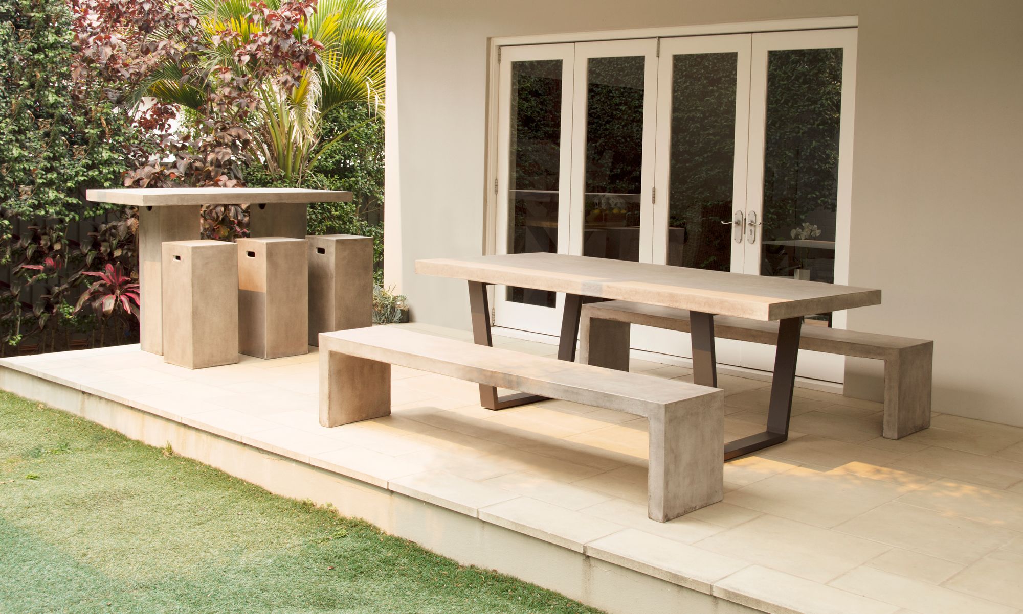 Remarkable Outdoor Living: Zen Outdoor Dining table with Aluminium Trapezoid shade Leg.  A contemporary outdoor dining set, featuring a concrete table and benches, is set against a lush garden backdrop, offering a harmonious blend of man-made structure and natural beauty.