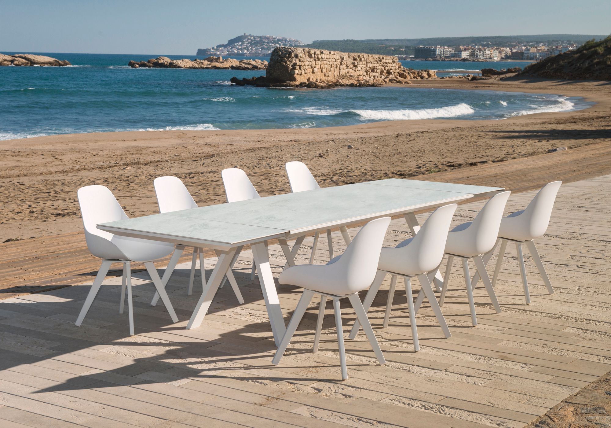Remarkable Outdoor Living: Neverland Outdoor Ceramic Extension Dining Table. A modern outdoor dining setting with a long table and chairs on a wooden boardwalk by the beach, overlooking the sea and coastline in the distance.