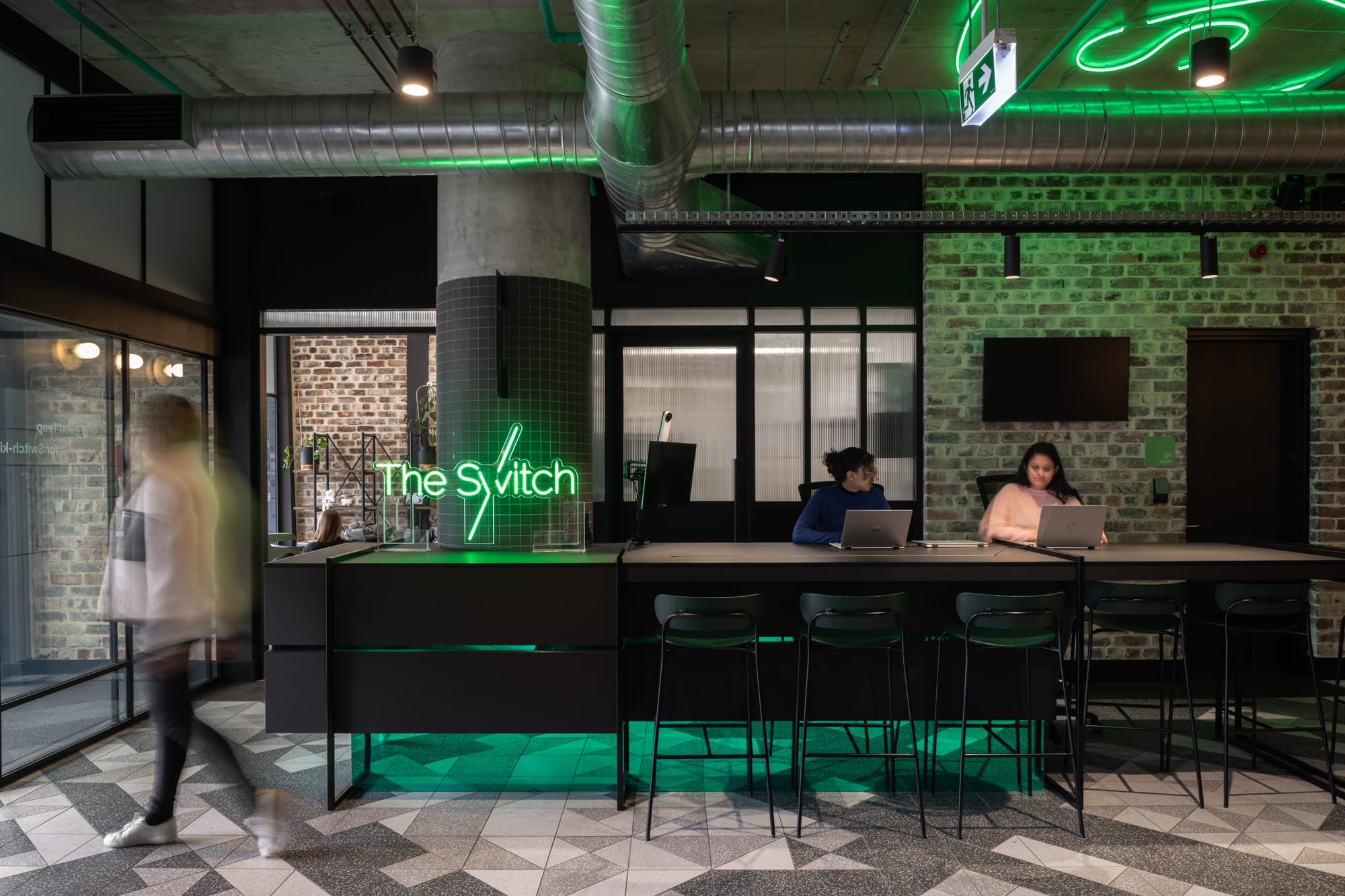 Thinking Differently About a Home Gives You Affordable ChoicesAn urban workspace with a green color theme, highlighted by a neon sign saying "The Switch." People are working at bar-style seating against a backdrop of green-painted brick walls and industrial-style lighting.