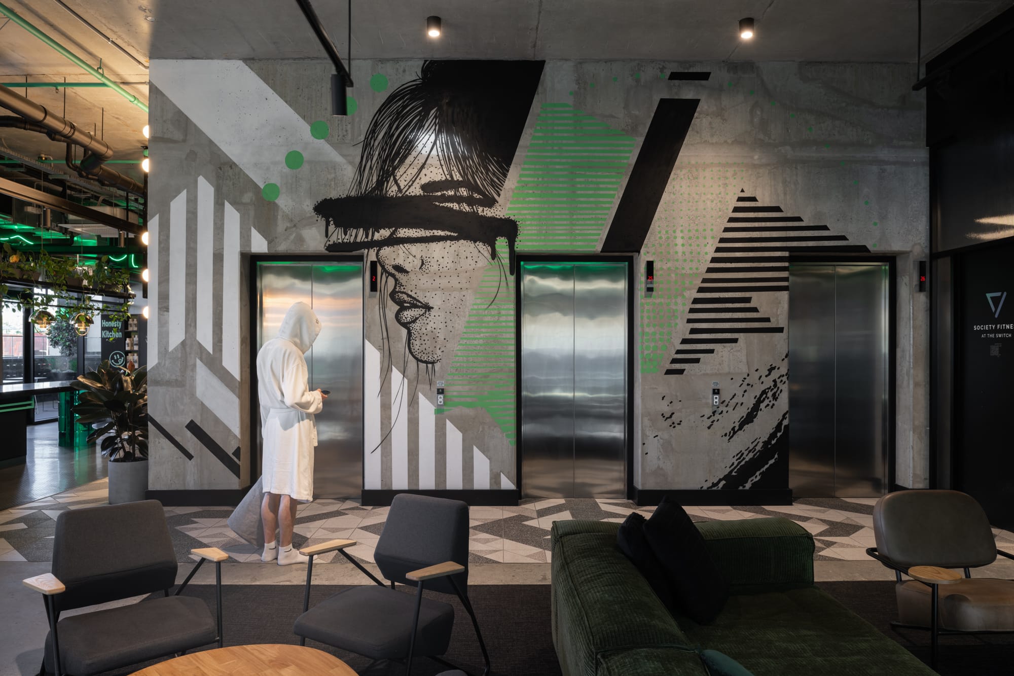 Thinking Differently About a Home Gives You Affordable ChoicesA modern building lobby with concrete floors and a large wall mural of a woman's face. A figure in a white robe stands looking at the mural, and plush green seating areas are scattered throughout the space.