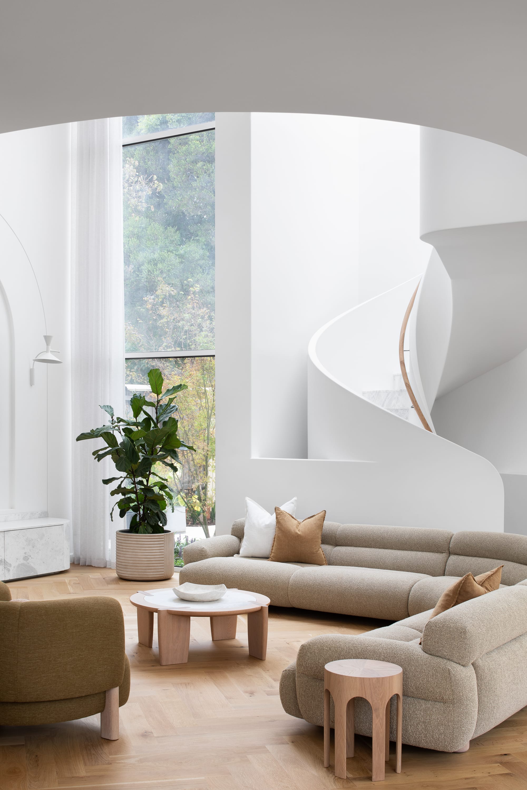 The Grove by Taouk Architects.A bright and airy living space with tall white walls and a spiral staircase with a sleek white balustrade. Natural light floods in from large windows overlooking greenery. The room is furnished with a beige sofa, matching armchairs, and a small wooden coffee table, creating a cozy yet modern atmosphere.