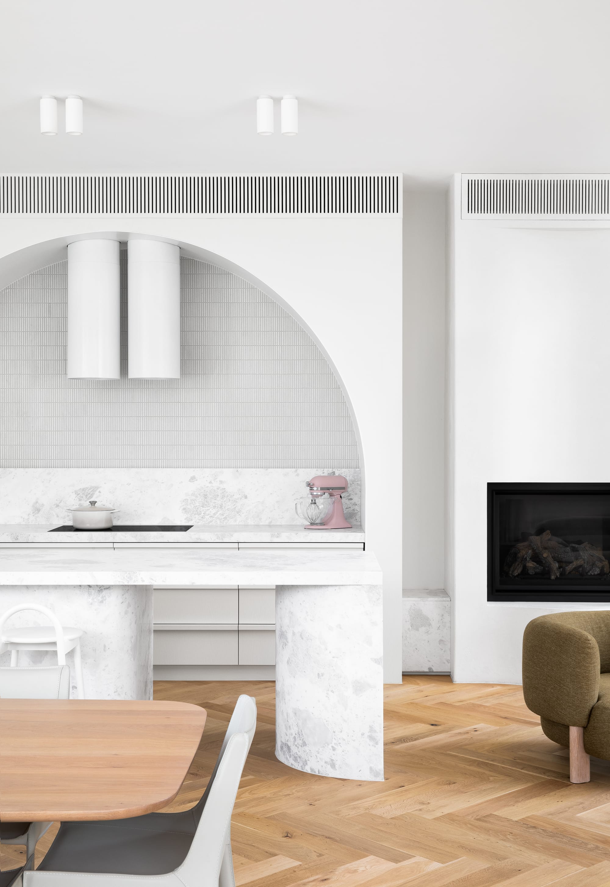 The Grove by Taouk Architects.A minimalist kitchen interior with a white marble island and a built-in cooktop under an arching range hood set against a tiled backsplash. To the right is a living area with a modern black fireplace. The space features herringbone wood flooring, white chairs around a wooden dining table, and a plush green armchair. The clean lines and white color palette give the room an open, serene feel.