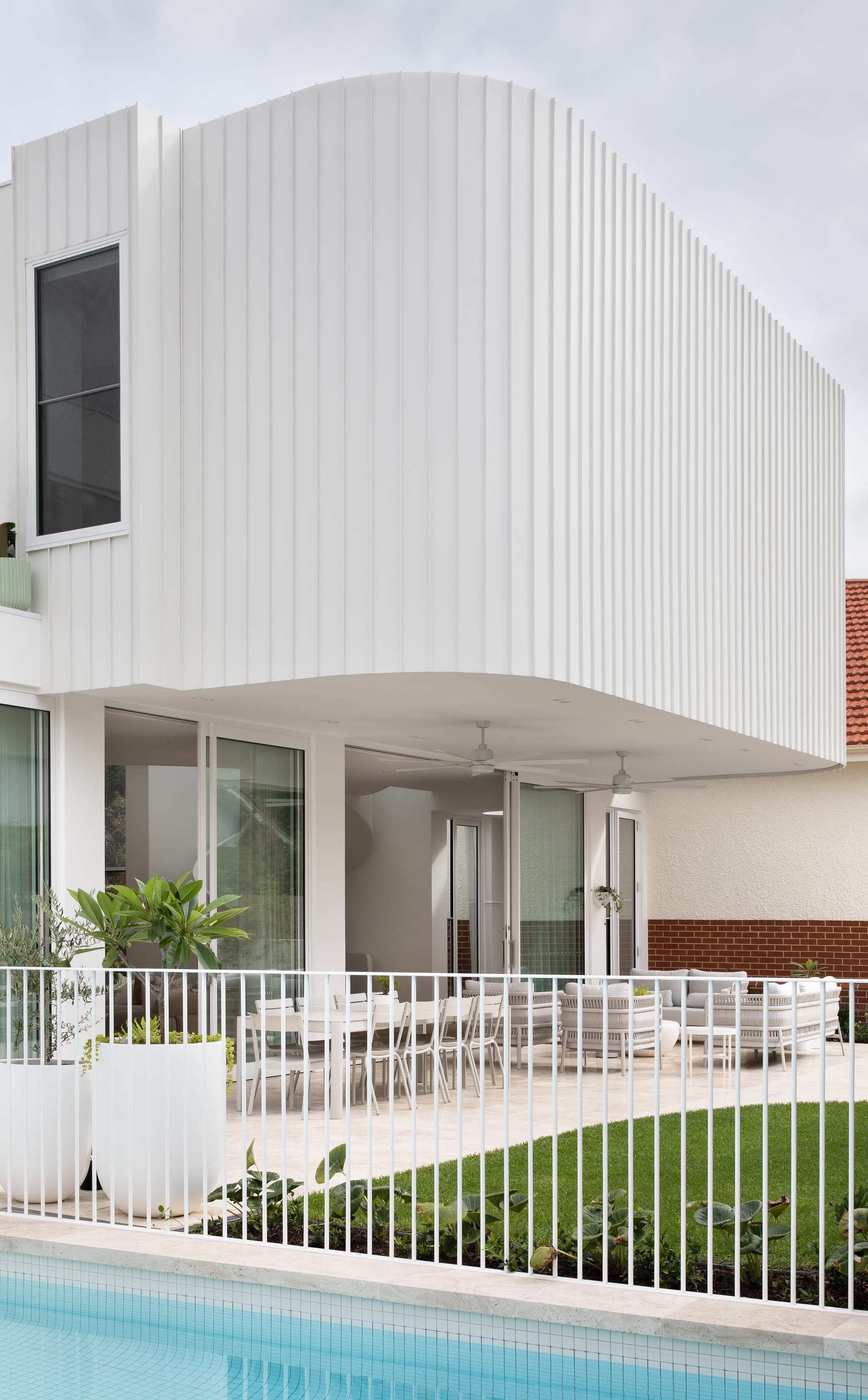 The Grove by Taouk Architects. A modern two-story house with a flat roof design and minimalist white facade. The ground floor features large glass windows and sliding doors that lead to a tiled patio with a swimming pool. A neat lawn and a white metal fence border the property.