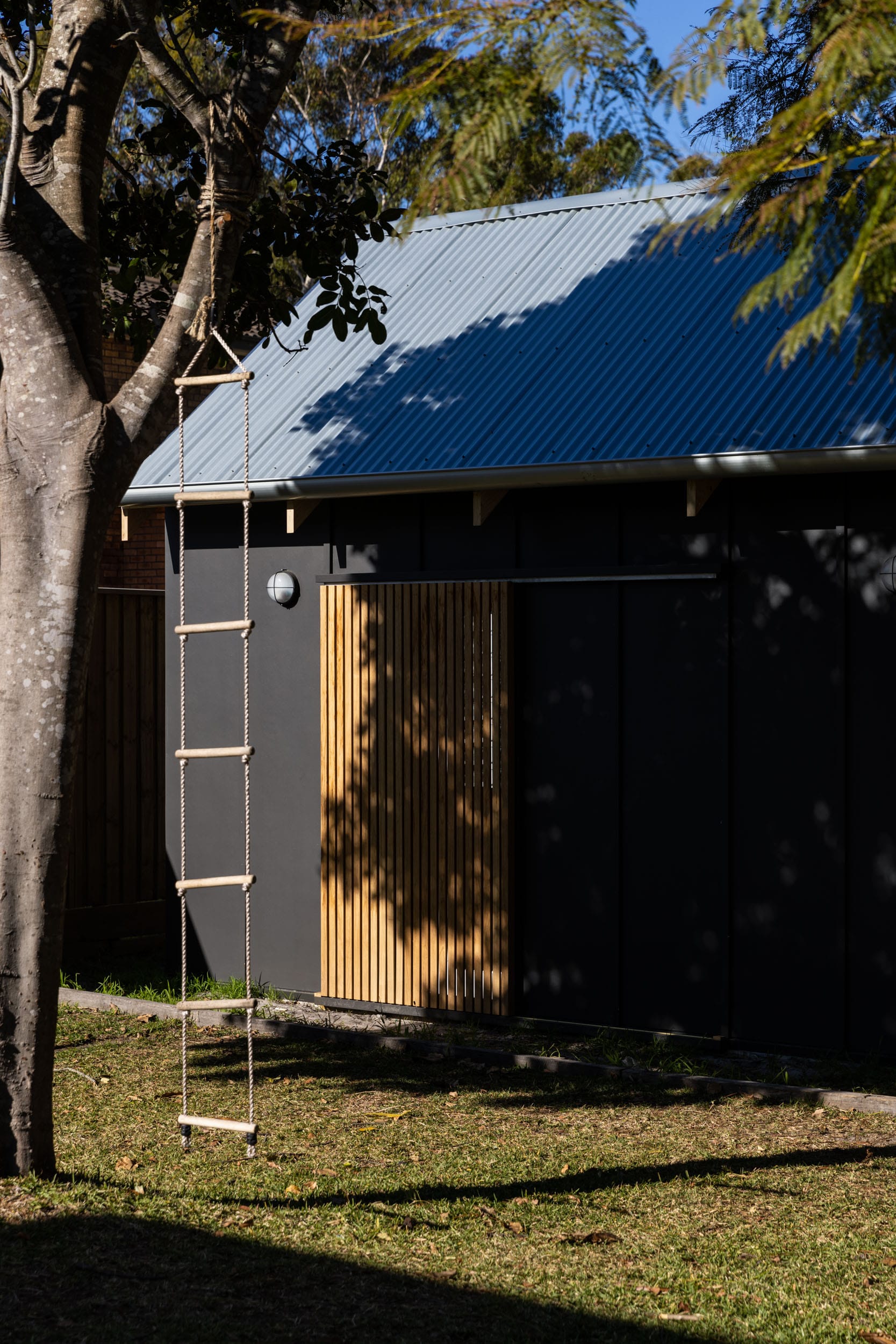 Drip-Dry House by Marker Architecture & Design. A shot of the yard with a rope ladder hanging from a tree and the read elevation of the dark cladding of the beach house