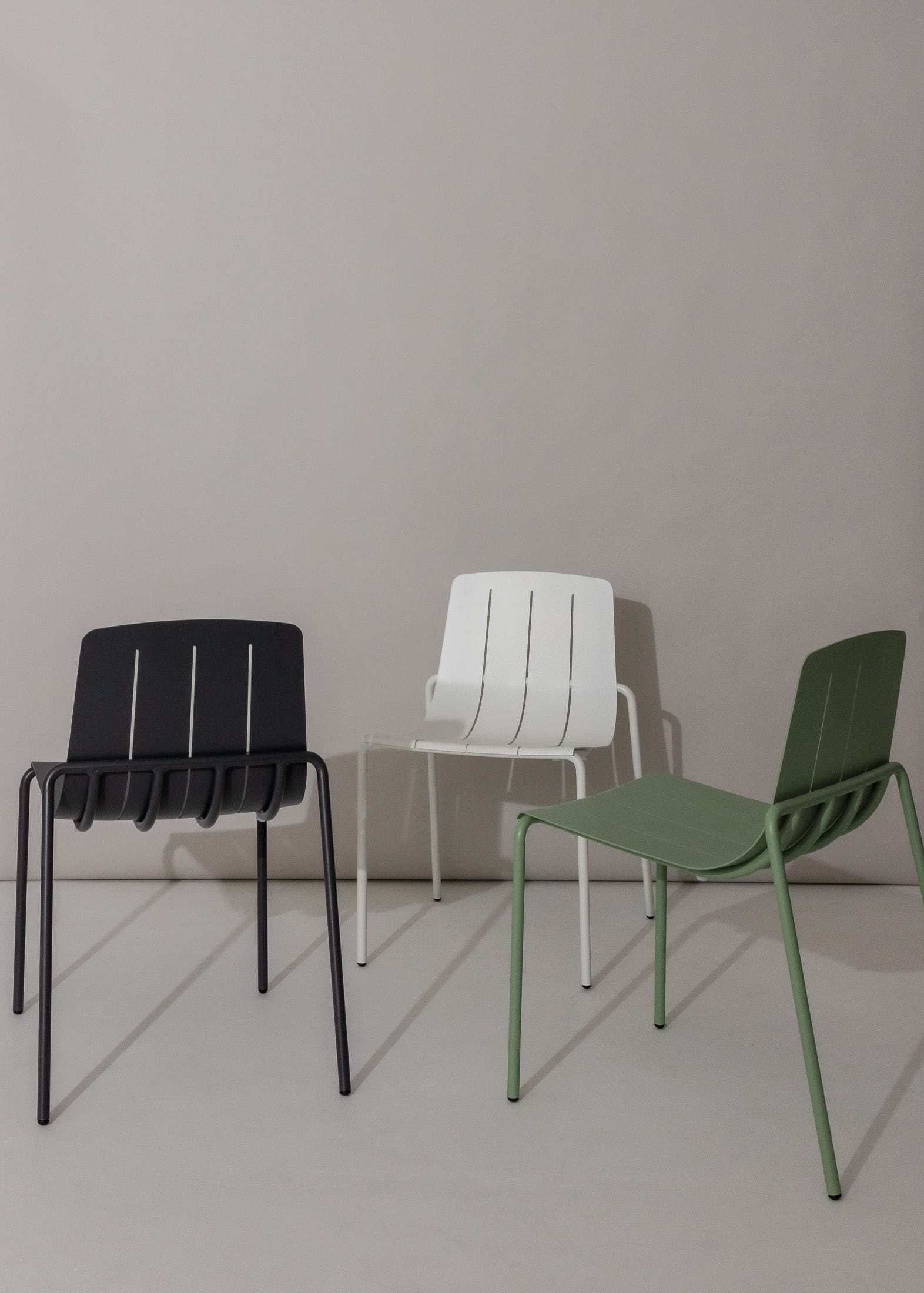 A trio of sleek outdoor chairs in a minimalist setting against a neutral background. The chairs have a modern design, with the first being black, the second white, and the third in a muted green, all with slender metal legs.
